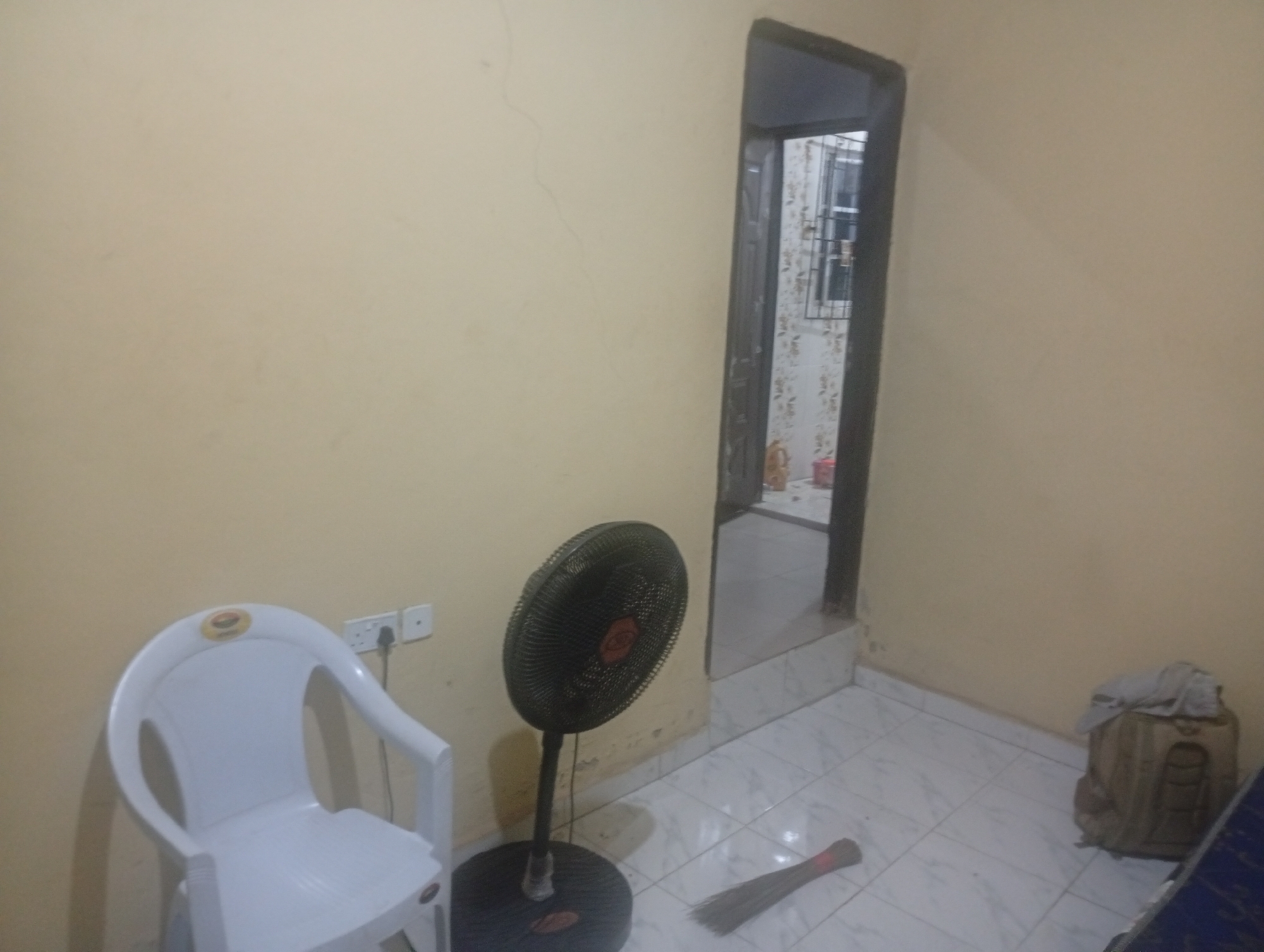 The image shows an interior scene within a room. On the left side, there is a white plastic chair with armrests, positioned against a light yellow wall. A white electrical outlet with a black cord plugged into it is mounted on the wall behind the chair. To the right of the chair, a black oscillating fan stands on a circular base. A broom with a red band around its bristles lies on the tiled floor in front of the fan. Further to the right, a beige backpack is placed on the floor, with a light-colored piece of fabric draped over its top. An open doorway is visible in the background, leading to another area with a window that has a metal grate. The floor throughout the room is tiled with a white and grey checkered pattern. The scene appears to be a residential interior. The specific location in Ekosa, Nigeria, is not determinable from the visual information provided.