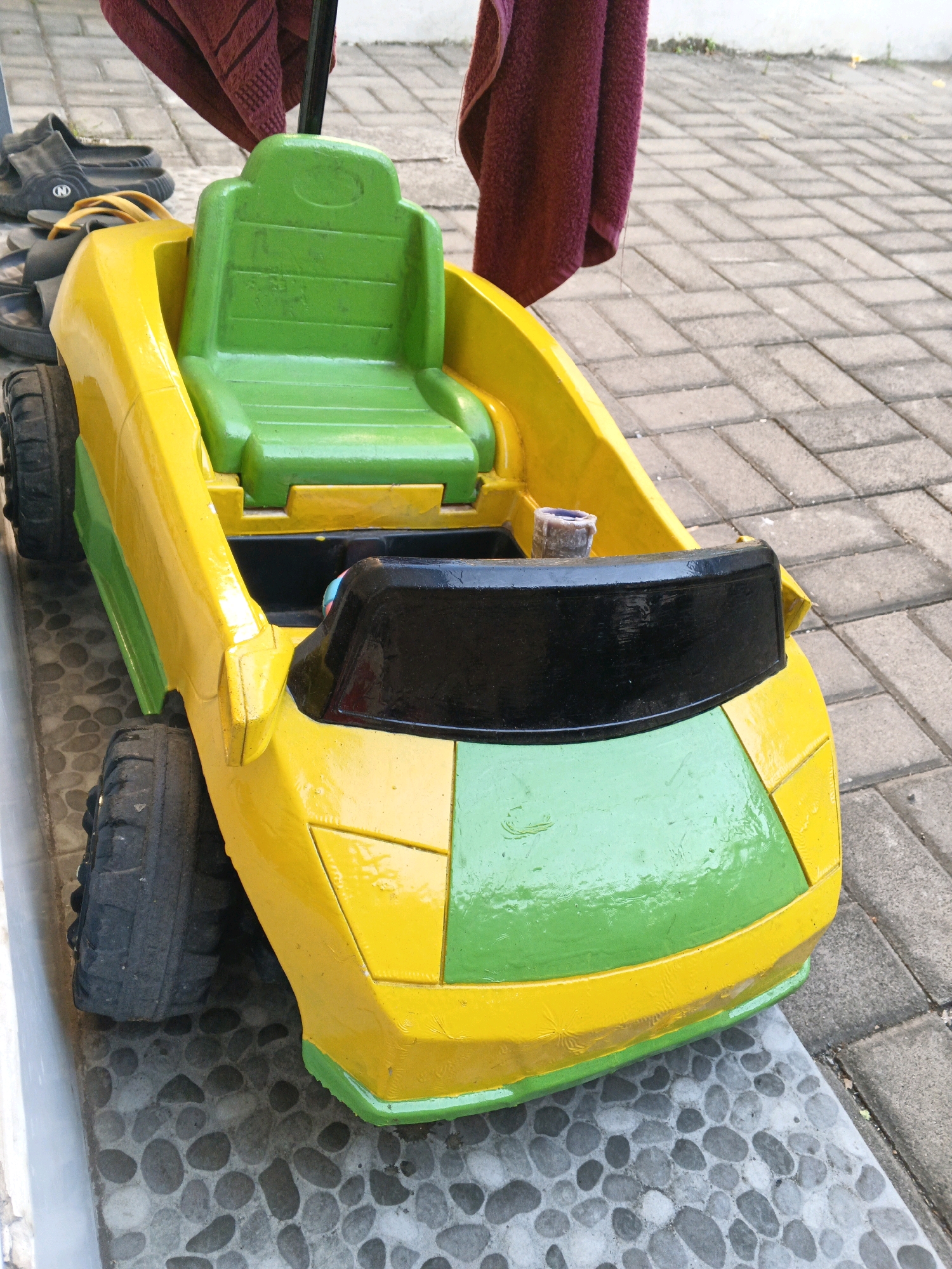A yellow and green toy car with black trim and wheels is parked on a grey and white tiled surface. The car has a green seat and a black windshield. A dark red towel hangs on a pole behind the car. To the left of the car are a pair of black sandals and another pair of dark colored sandals. The ground is made of grey and white rectangular tiles arranged in a herringbone pattern, with round, grey pebbles bordering the tiled area. The scene appears to be outdoors, possibly in a residential area in Denpasar, Indonesia.