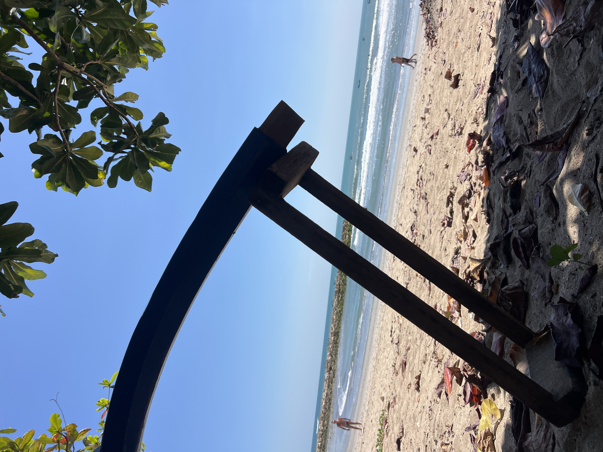 This image shows a tropical beach scene in Kuta, Indonesia. In the foreground, a wooden structure, possibly part of a shelter or seating, is angled across the sand. Fallen leaves and some greenery are scattered on the sand. Beyond the foreground, a sandy beach stretches towards the ocean, with gentle waves breaking on the shore. A line of rocks creates a barrier in the shallow water. In the distance, two men are walking on the beach, and several small boats are visible on the water. The sky is clear and blue, with green foliage from overhead trees framing the top of the image. The scene depicts a sunny day at the beach, with a relaxed atmosphere.