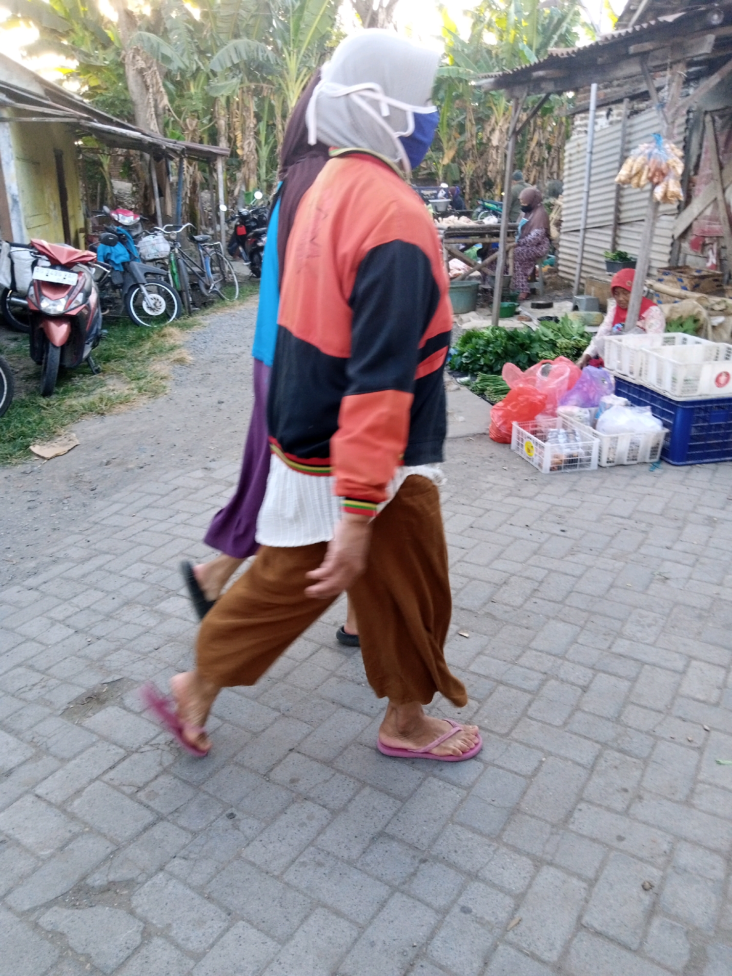 In Sono Ageng, Indonesia, a street scene unfolds with a focus on two individuals walking on a paved walkway. The person in the foreground, wearing a light grey hijab, blue face mask, and an orange and black jacket over brown pants, walks away from the camera. Their right foot is visible, clad in a pink flip-flop. To their left, another person walks in the same direction, partially obscured, wearing a purple garment and black flip-flops. The environment includes several motorcycles parked on a dirt path to the left, with one red scooter prominently displayed. To the right, a market stall area is visible, with wooden structures, corrugated metal roofing, and displays of produce, plastic bags, and crates. In the background, lush green vegetation, including banana trees, suggests a tropical setting. The lighting indicates it is daytime.