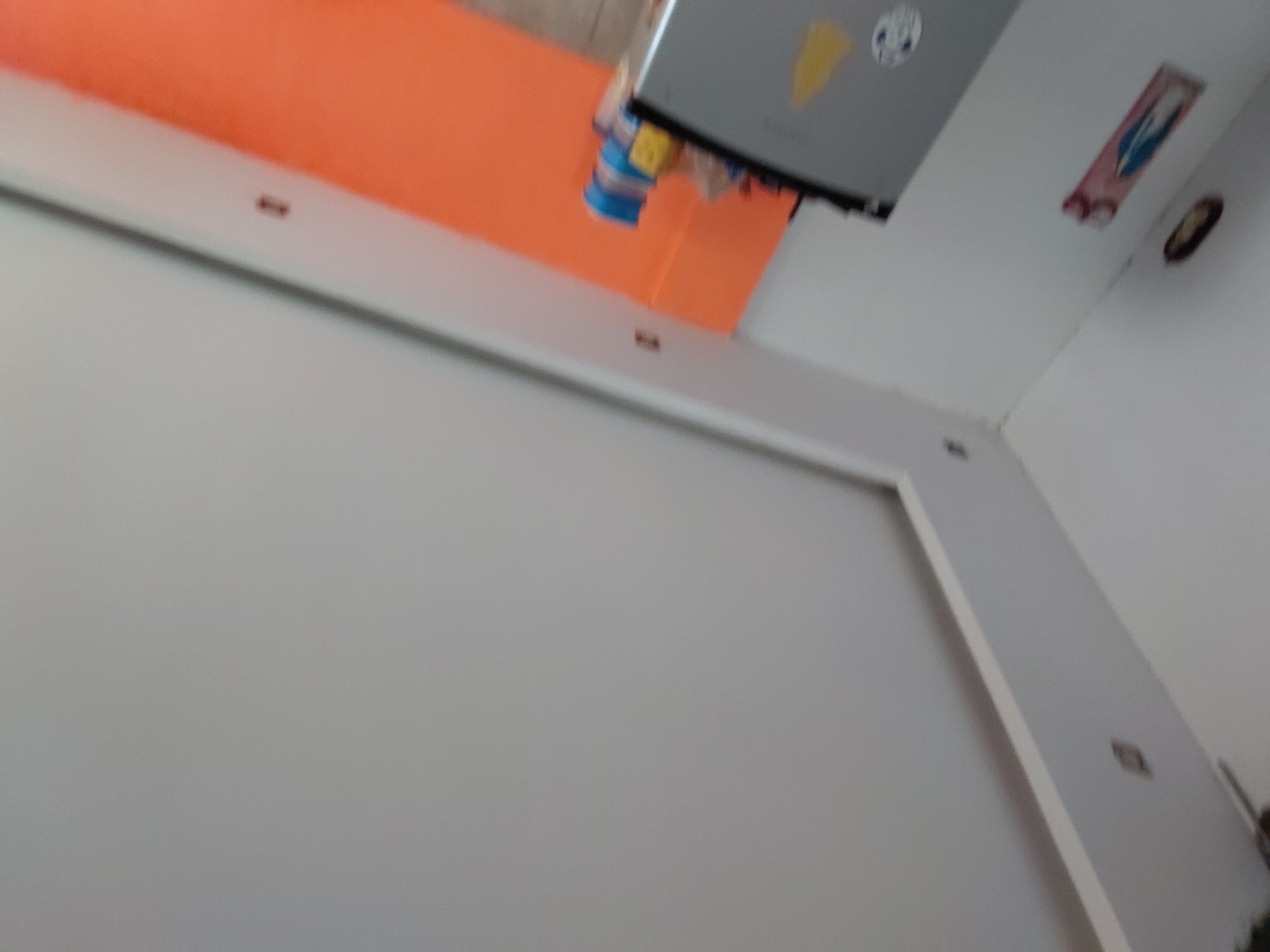 The image captures an indoor scene, viewed from a low angle looking upwards towards the ceiling and upper walls. The perspective is tilted, suggesting a handheld shot.
The primary subjects are the interior architecture and mounted fixtures. Walls are visible, one painted a distinct orange color, while others and the ceiling are white or light gray. White architectural trim or beams frame sections of the walls and ceiling.
High up, near the junction of the orange wall and the ceiling, a gray or silver rectangular device is mounted. This device, possibly an air conditioner unit or projector, has a yellow triangular sticker and a white circular logo on its underside. Below this device, several blue and yellow objects appear to be hanging or attached, their exact nature unclear but they resemble small containers or decorative items.
On an adjacent white wall or ceiling section, a small, dark rectangular object is visible, displaying some blue and red colors, possibly a religious icon or a small picture. Further along the white wall/ceiling, a dark oval-shaped fixture, possibly a light or vent, is present. Small dark specks or marks, which could be fasteners or minor imperfections, are visible on the white trim and surfaces. Another small, dark square object is embedded in a white beam, possibly a switch or outlet cover.
There are no people present, and no discernible activity or event is taking place. The time of day is not indicated, but the space appears well-lit, likely by artificial lighting or bright natural light from an unseen source. No visible text is legible in the image, as details are somewhat blurry. The scene depicts a static interior space.