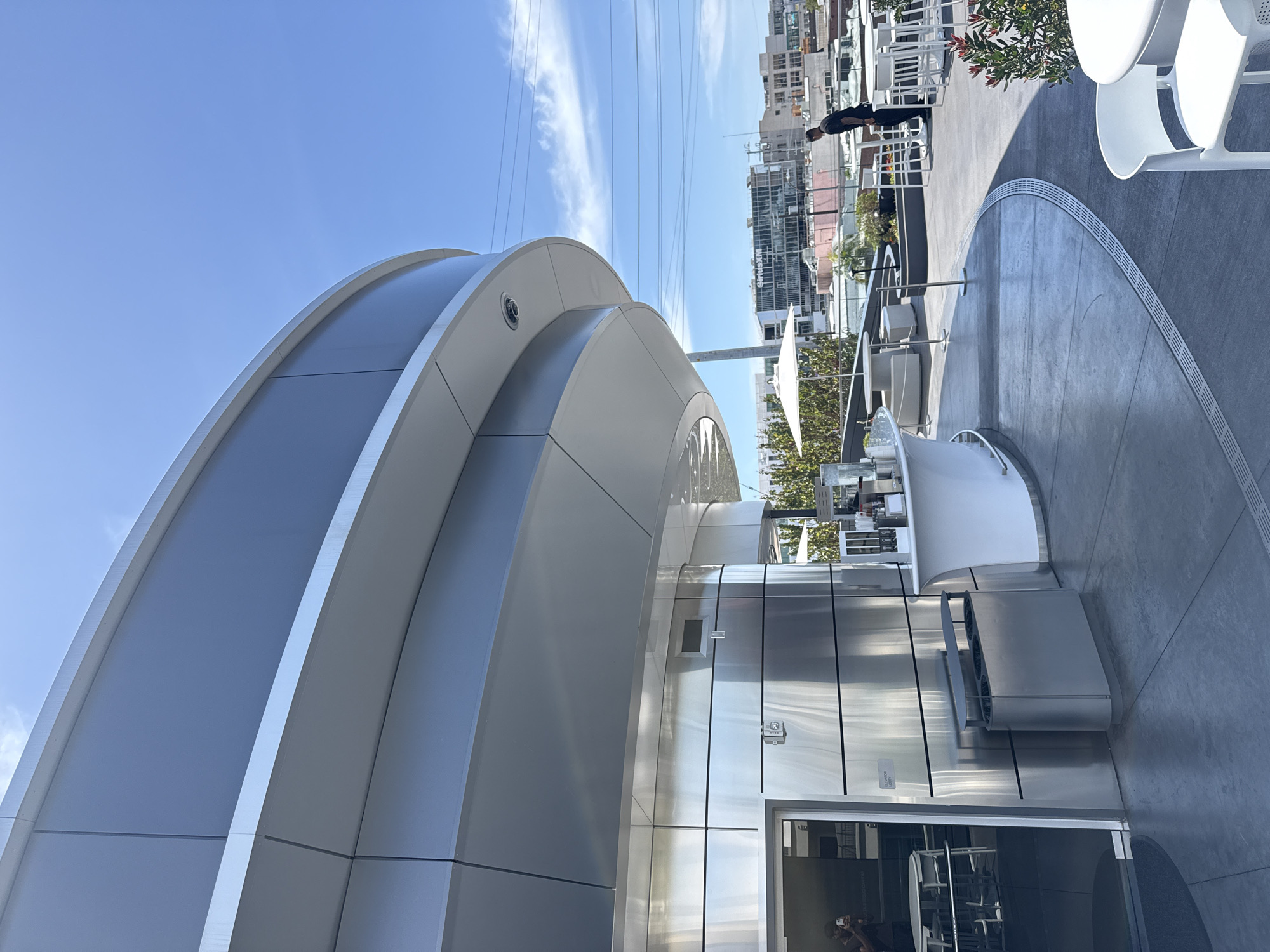 The image depicts a modern architectural structure with a curved, layered exterior primarily in shades of gray and silver. The structure appears to be located in an urban environment, as taller buildings are visible in the background. In the background, a person is standing on a rooftop or elevated area with white patio furniture. There are power lines visible against a partly cloudy blue sky. The structure features reflective surfaces, suggesting the use of metal or similar materials. The presence of an entrance suggests it is a building or facility. The architectural style is modern and sleek. The image appears to be taken at ground level, looking upwards towards the building. Based on its appearance in the photograph, the building is located in the city of West Hollywood, United States of America.