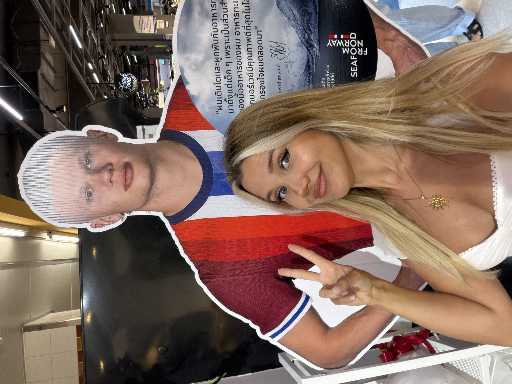 In Patong, Thailand, a woman with blonde hair and a sun-shaped necklace poses for a selfie in front of a life-size cardboard cutout of a man with blonde hair. She is wearing a white top and is making a peace sign with her right hand. The cutout is wearing a red, white, and blue striped shirt. To the right of the cutout, there is a circular sign with images of mountains and text in Thai and English that reads "SEAFOOD FROM NORWAY". The background shows a supermarket environment with fluorescent lighting and shelves.