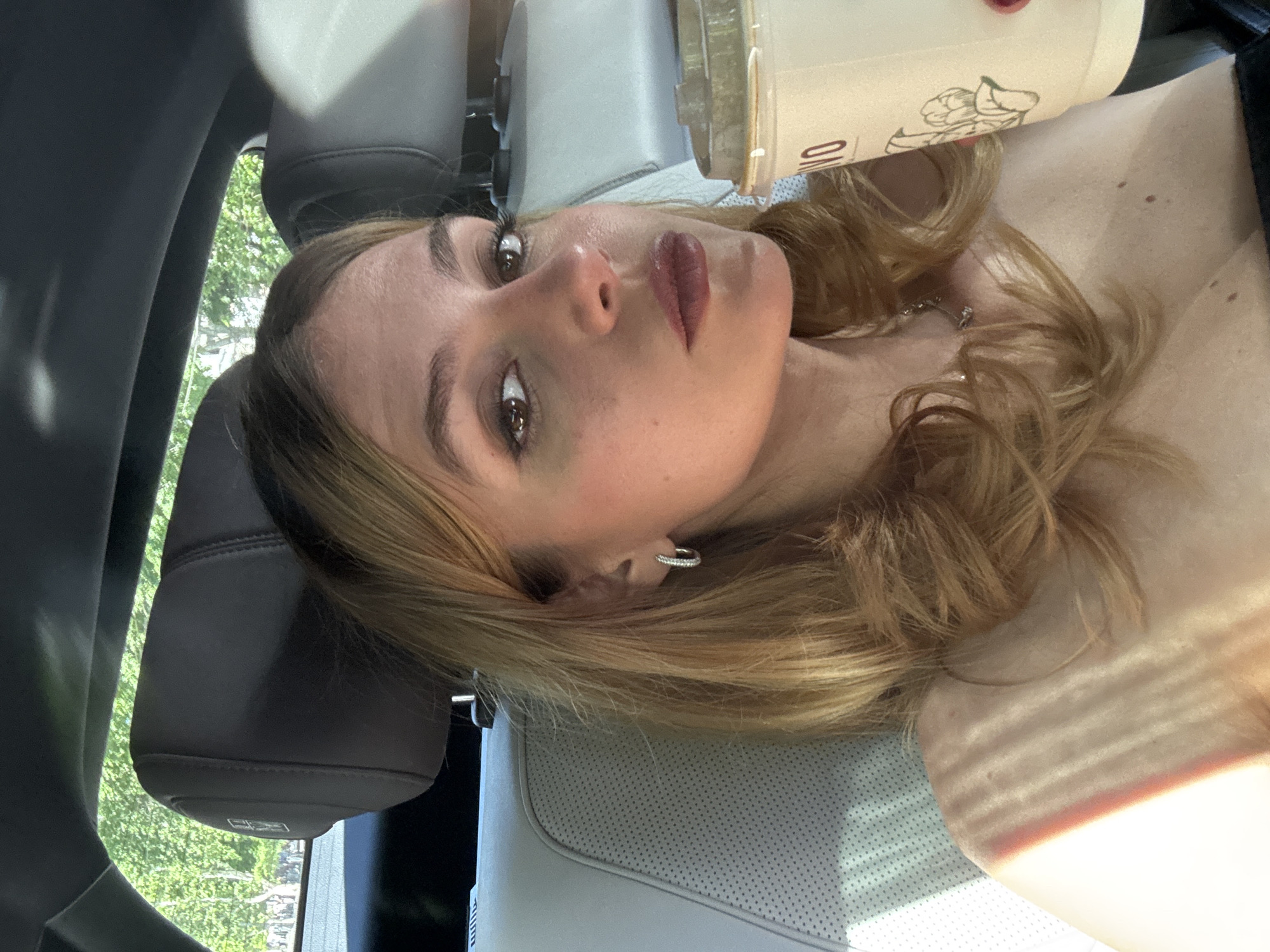 A medium shot depicts a woman with light brown hair, light skin, and a neutral expression. She is positioned in a car, possibly a passenger seat, as indicated by the headrest and the seat visible. She is holding a drink container, with a design visible on it. The environment outside the car shows greenery, possibly trees, suggesting an urban or suburban setting. There are no specific details to pinpoint the location to Yerevan, Armenia. The scene is casual and unposed.