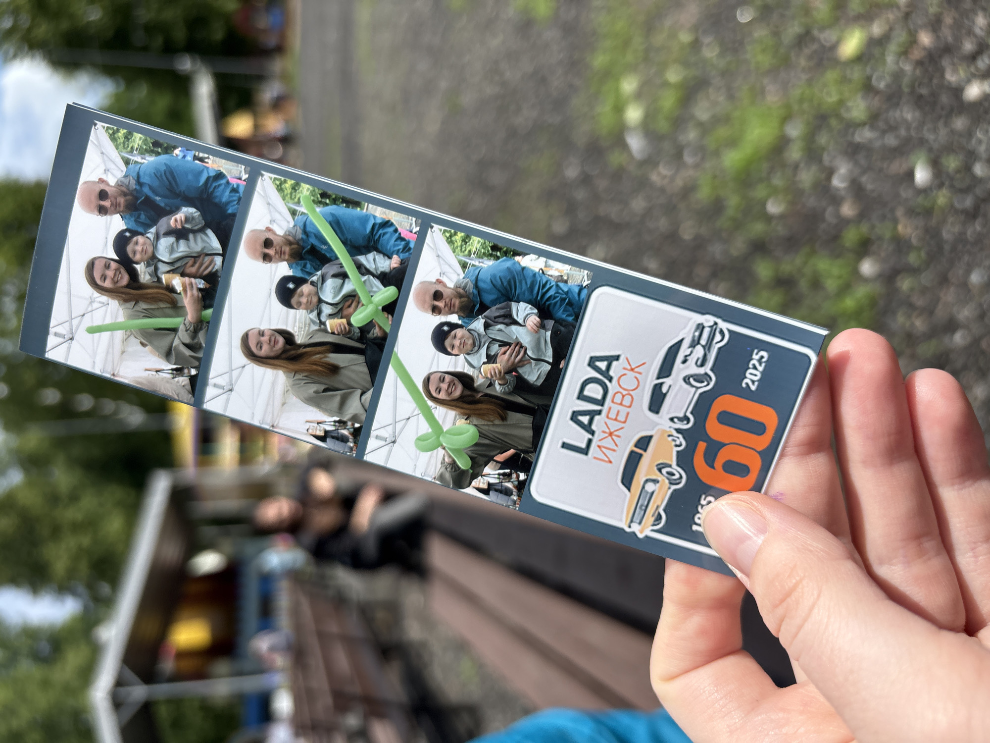 A person holds a photo booth strip in their hand. The photo strip contains three identical pictures of a woman, a bald man wearing sunglasses, and a child. The woman holds a green balloon animal. The strip also contains text in Russian and English, and illustrations of two Lada automobiles, a 1965 and 2025 model. The text reads "LADA ИЖЕВСК" and "60 1965 2025".
The background is out of focus, but appears to be an outdoor setting with trees, and buildings. It is plausible that the photo was taken in Izhevsk, Russia, given the information on the photo booth strip.