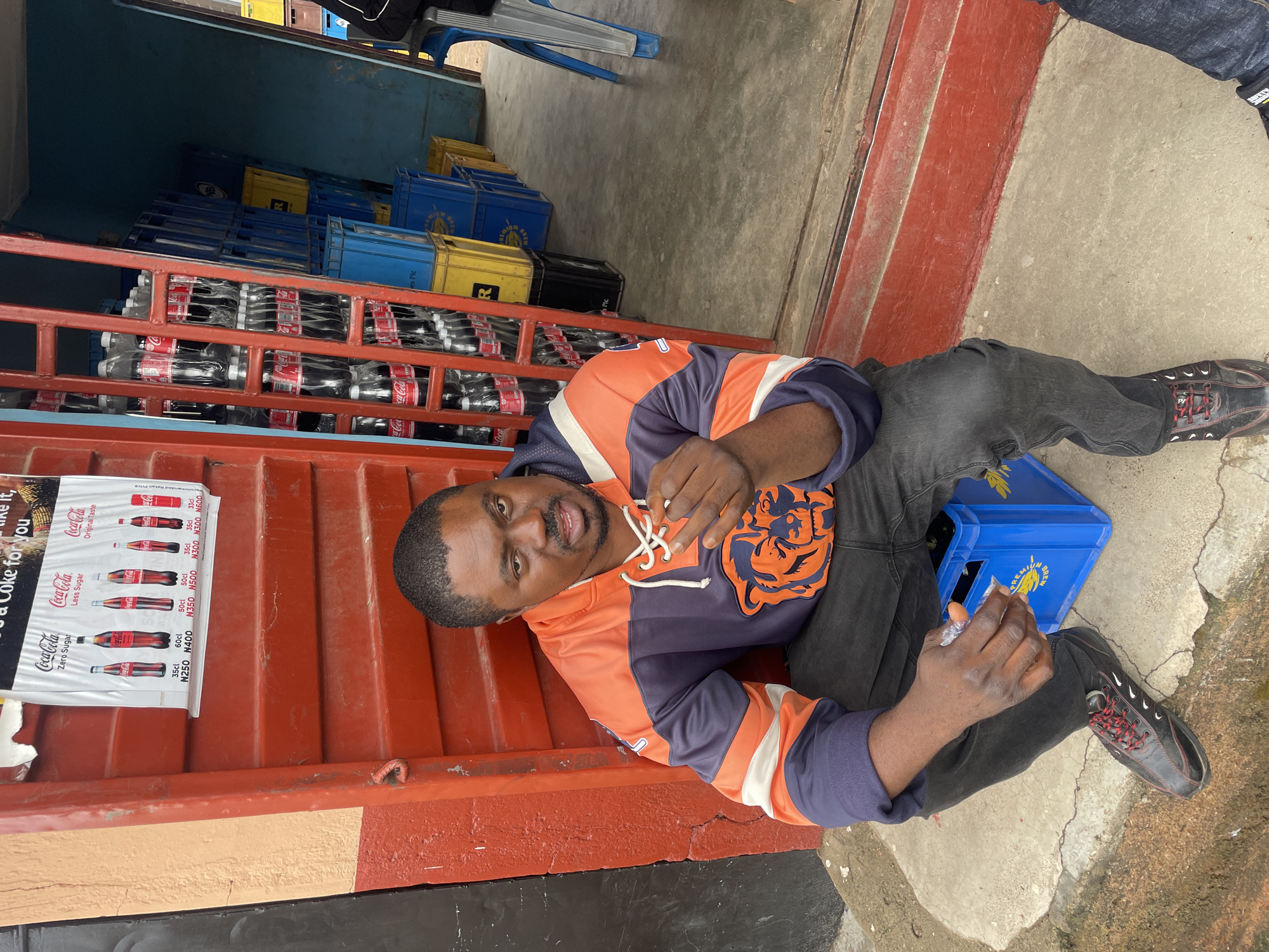 A man sits on a blue plastic crate in front of a storefront in Jos, Nigeria. The man wears an orange and navy hoodie with an animal emblem on the chest, dark pants, and black shoes with red laces. He holds a small, translucent plastic bag in his hands. Behind him, stacked on a red metal rack, are rows of Coca-Cola bottles in clear plastic packaging. A Coca-Cola advertisement is affixed to the red metal rack. Yellow and blue crates are stacked inside the storefront. The scene is illuminated by daylight.