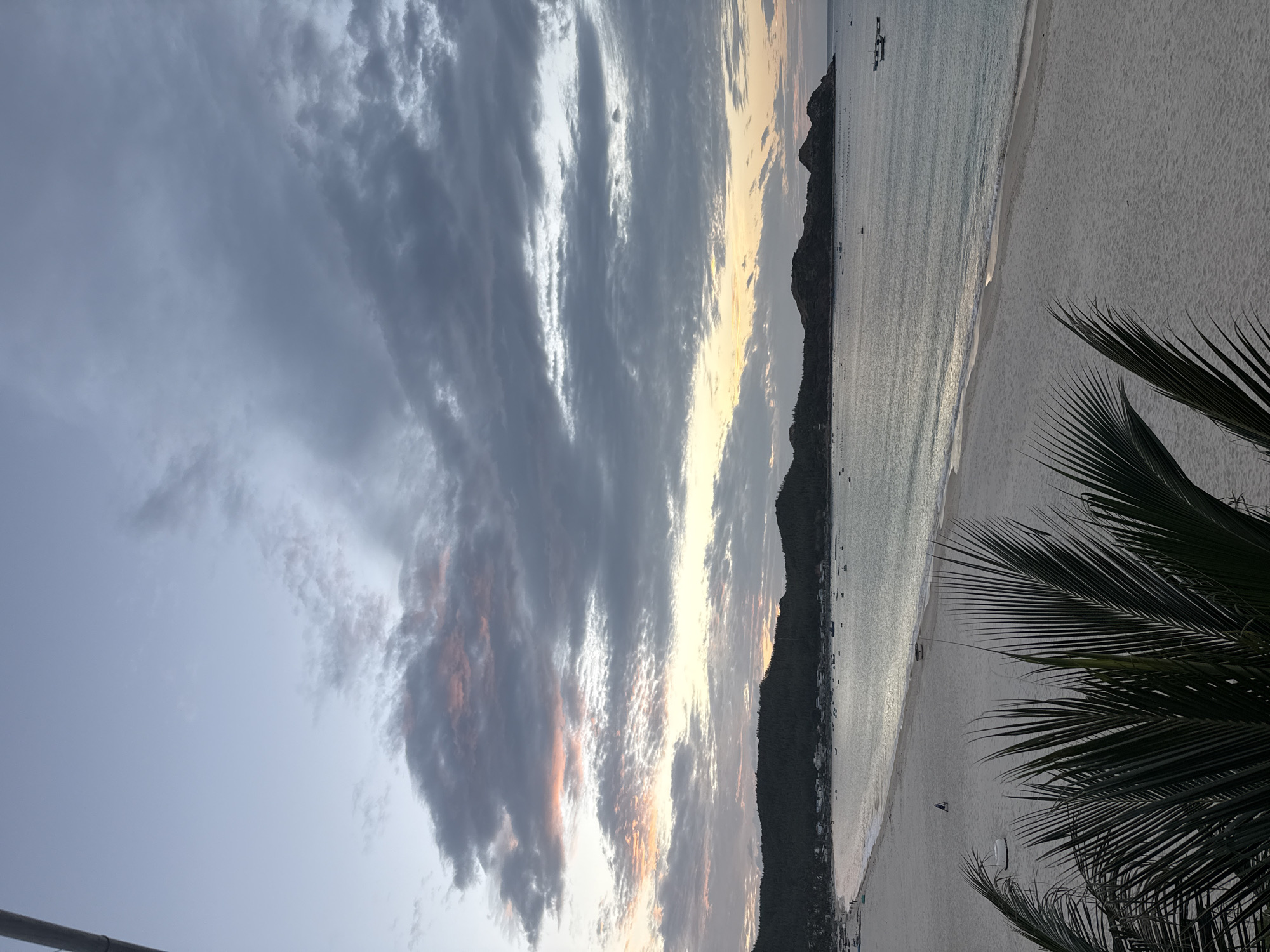 This image depicts a wide-angle view of a tranquil beach scene at dusk, likely in Song Cau, Vietnam. The foreground is dominated by the lush, dark green fronds of palm trees, obscuring the bottom portion of the frame. Beyond the foliage, a vast expanse of pale sand stretches out, meeting the gentle waves of the ocean. A solitary figure can be seen walking on the beach in the mid-ground, and a couple of white beach chairs or small boats are also visible on the sand. The sea is calm, reflecting the subdued light of the sky, and dotted with numerous small boats scattered across its surface. In the distance, a chain of dark, tree-covered hills forms a dramatic backdrop to the coastline, with a small cluster of buildings visible nestled at the base of the hills. The sky is a canvas of soft, muted colors, transitioning from a pale blue at the top to hues of yellow and soft pink near the horizon, with dramatic grey clouds streaked across the expanse. The overall atmosphere is serene and peaceful, capturing the quiet beauty of the coastline at twilight.