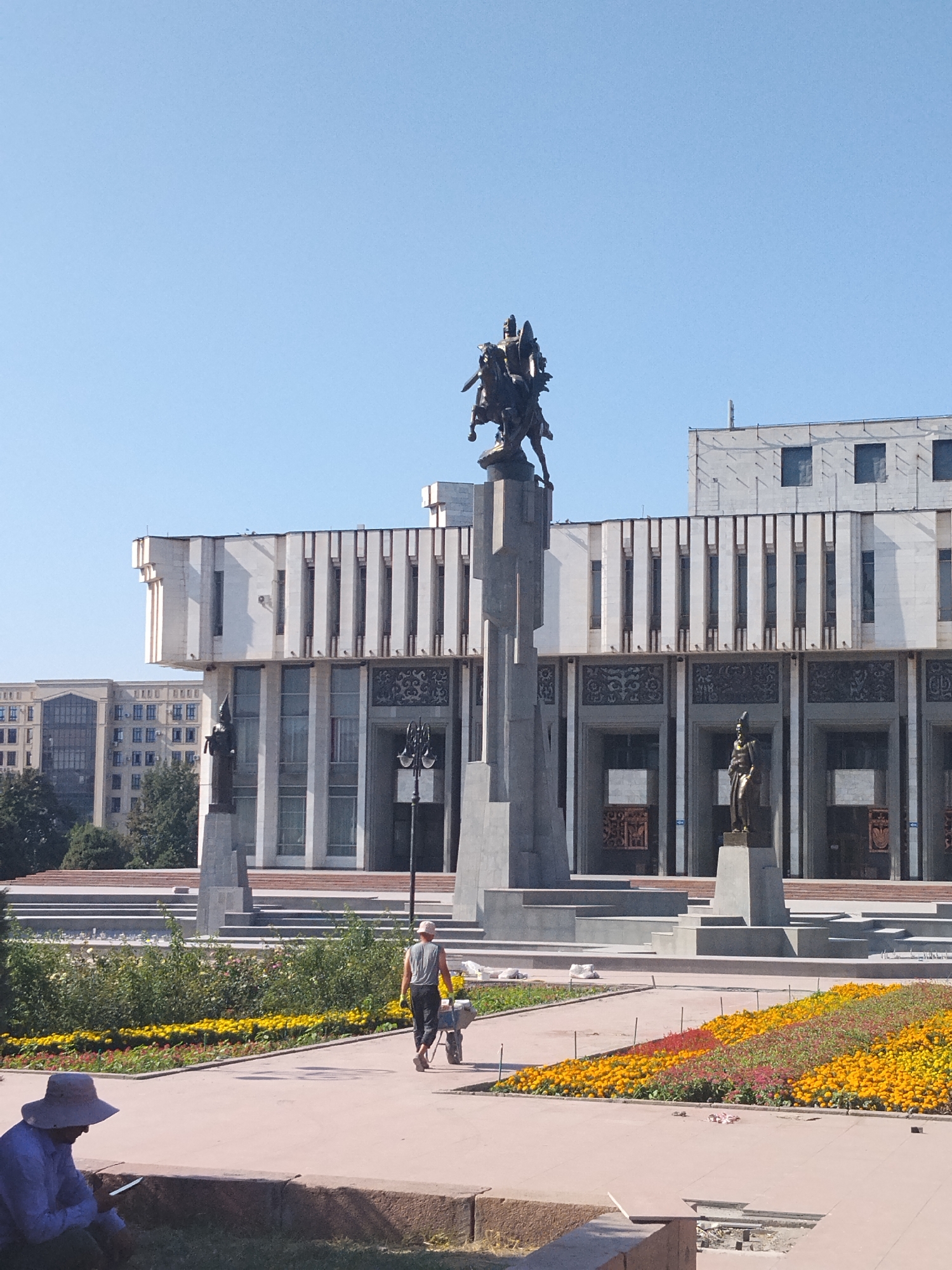 In front of a large, modern building with a colonnade of vertical fins and carved decorative panels, a bronze equestrian statue stands on a tall, tiered pedestal. To the left of the equestrian statue, a smaller bronze statue of a standing figure is visible. In the foreground, a man wearing a hat and a blue shirt is sitting and looking at a mobile phone. Further into the scene, a man wearing a hat and a grey tank top is pushing a wheelbarrow filled with yellow flowers across a paved walkway. A flowerbed with rows of yellow, red, and pink flowers runs along the walkway. The background shows more modern buildings and a clear blue sky. This scene is located at Ala-Too Square in Bishkek City, Kyrgyzstan.