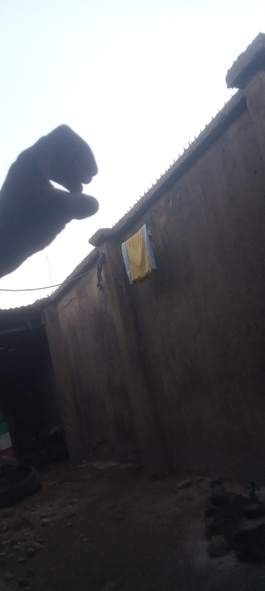 The scene depicts the exterior of a mud building in Yelwa, Nigeria. A shadowed hand is visible in the foreground. The building has a rough, textured wall and a roof line with pointed projections. A yellow and blue cloth hangs on the wall. The ground is uneven. The lighting suggests early morning or late afternoon.