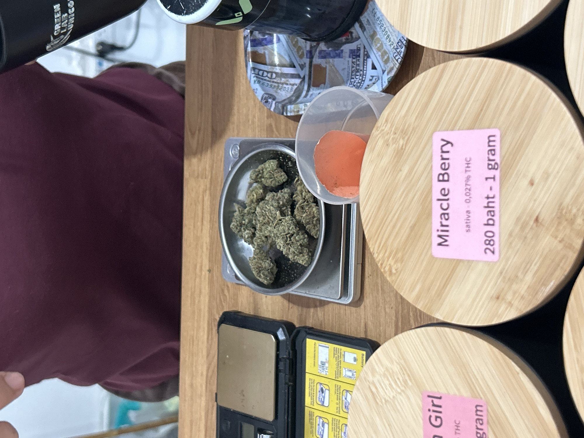 The image shows a close-up view of a wooden counter in what appears to be a retail establishment in Ratsada, Thailand. On the counter, there are several items related to the sale of cannabis. In the center, a small digital scale holds a pile of dried cannabis buds in a metal weighing dish. To the left of the scale is a black digital scale with an open lid, displaying instructions in diagrams. To the right of the weighing dish is a small, clear plastic cup containing a small amount of orange powder. Behind this, a tray printed with US dollar bills is visible, partially obscured by a black cylindrical object with a logo that reads "GREEN LAB". In the foreground, two wooden circular containers with bamboo lids are prominently featured. Pink labels are affixed to the lids: one reads "Virgin Girl" and "grams THC", while the other reads "Miracle Berry", "sativa - 0,027% THC", and "280 baht - 1 gram". A person's hand and arm, wearing a dark maroon shirt, are visible at the top of the image, suggesting they are interacting with the items on the counter. The environment appears to be a dimly lit retail space.