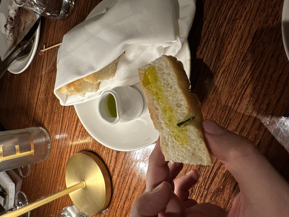 The image captures an indoor dining scene, specified as taking place in the city of London, United Kingdom. A person's left hand, visible in the lower central and left foreground, holds a rectangular piece of focaccia bread, which is generously drizzled with olive oil. The bread exhibits a light brown crust and a pale, airy crumb with visible air pockets. On a white ceramic plate directly behind the held bread, several other pieces of similar focaccia are partially wrapped in a white cloth napkin. Adjacent to the bread plate, a small white ceramic pitcher holds additional olive oil. To the left, a golden-colored lamp stand base is visible. In the upper background, a clear glass bottle with a golden "E" monogram is present, along with a white plate containing remnants of a dark, crumbly substance and a fork and knife, indicating a recently consumed dessert. A clear glass of water is also visible on the right. The entire scene is set on a polished reddish-brown wooden table with a visible grain, under warm, ambient lighting.