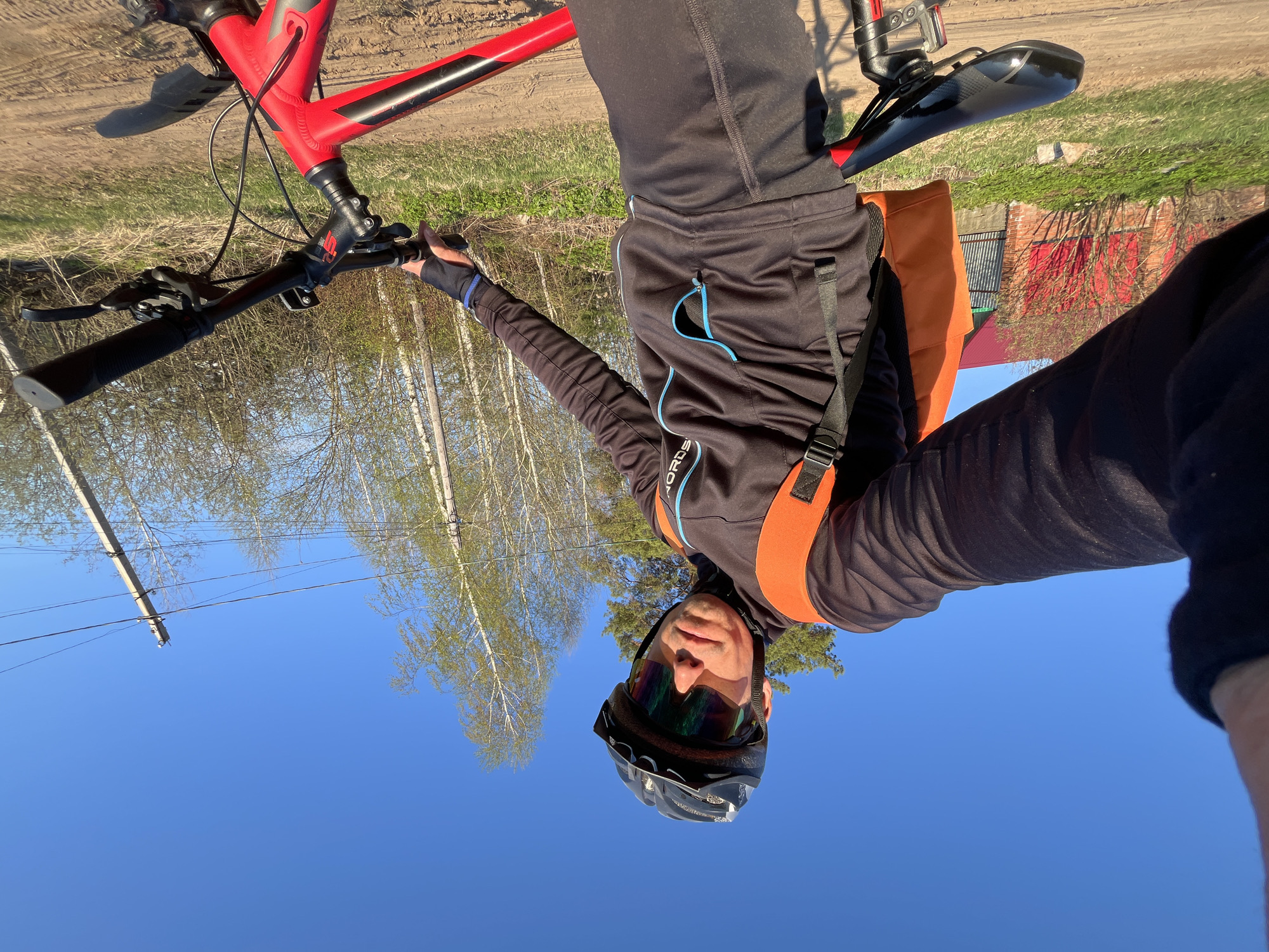 A man is taking a selfie while riding a red mountain bike on a dirt road. He wears a black helmet and sunglasses. He is wearing black athletic clothing with an orange backpack. The background includes birch trees, a blue sky, and a brick building with a red roof. A power pole and lines are visible in the sky. The location appears to be rural, possibly on the outskirts of a town or village. The specific location within Chaikovsky, Russia, is difficult to ascertain without additional location information in the image.