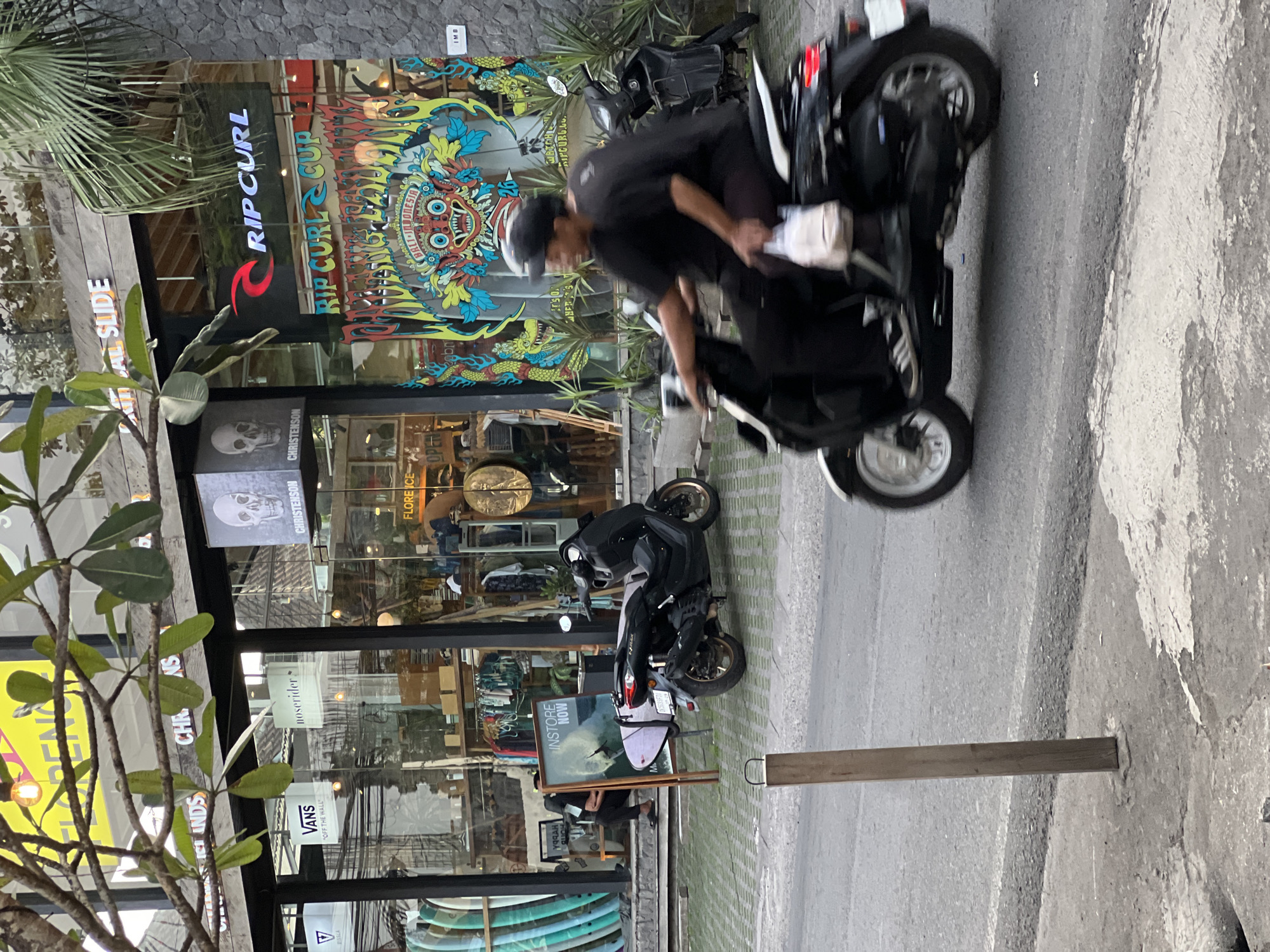 In Kuta Selatan, Indonesia, this image captures a street scene with several shops and a man riding a scooter. The storefronts display various retail brands including "VANS" and "RIP CURL," with surfboards visible outside one establishment. The Rip Curl store features a large, colorful mural depicting a traditional Balinese mask. A dark-colored scooter is in motion in the foreground, with a person wearing a black shirt and a helmet riding it. Another black scooter is parked to the left. The environment appears to be a commercial street with a sidewalk and paved road.