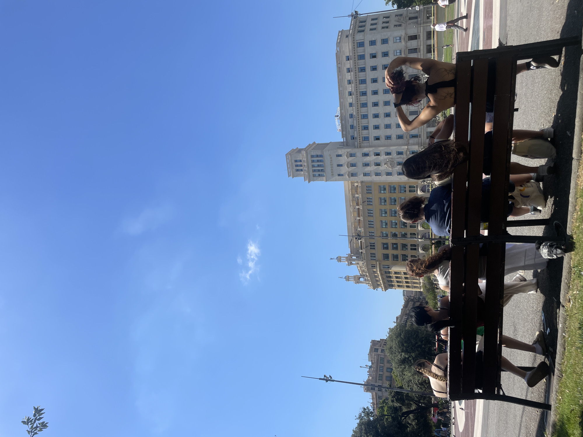 The image shows a group of young people sitting on a park bench on a sunny day. The scene is outdoors with a clear blue sky and some scattered clouds. In the background, there are several large, ornate buildings, suggesting an urban setting. The people on the bench appear to be relaxed and engaged in casual conversation or individual activities. One person is seen adjusting their hair, while others are looking down or away from the camera.
The time of day is likely midday or early afternoon, given the bright sunlight casting shadows. The weather seems pleasant and warm. The architecture of the buildings hints at a European city, and the provided context specifies Barcelona, Spain. The overall mood appears to be one of leisure and enjoyment of a sunny day. There is no visible text in the image.