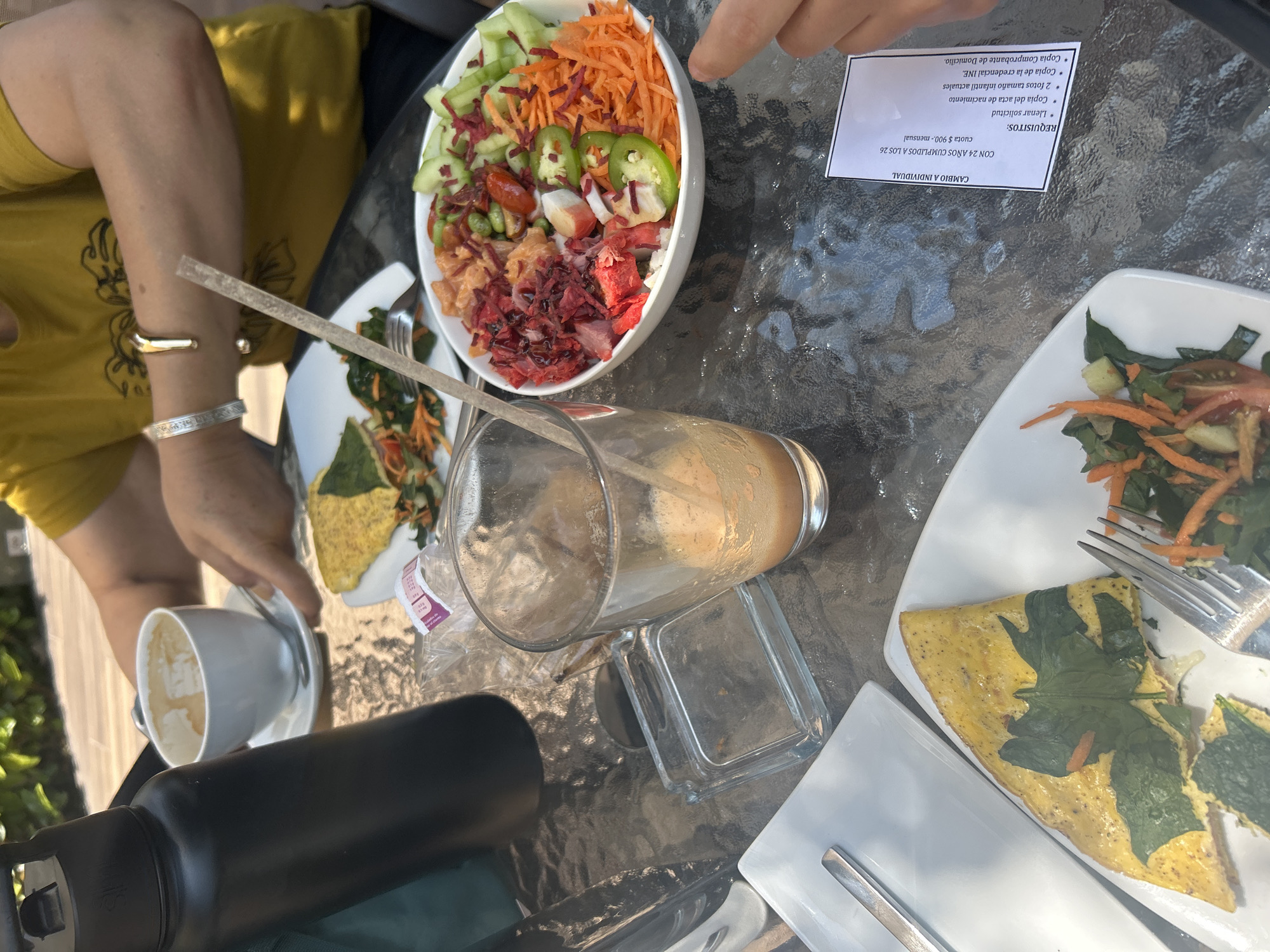 Here's a forensic description of the image:
**Setting:** An outdoor dining area, likely a patio or terrace. The tabletop is glass with a textured surface. The scene appears to be in San Bernardino Tlaxcalancingo, Mexico based on text present on the paperwork, although there is no verifiable information within the scene to confirm this.
**Objects:**
* A large white bowl containing a salad composed of mixed ingredients, including cucumbers, carrots, beetroot, jalapenos, and other red items.
* Two white plates, each with a portion of food that looks like an omelet with spinach and a side salad.
* A tall, empty glass with a straw inside.
* A small white cup, presumably containing coffee or a similar beverage.
* A black water bottle.
* Cutlery, including forks and knives on the plates.
* A clear, small square glass container.
* A piece of paper, possibly a document or receipt, with Spanish text containing the words "REQUISITOS", "CAMBIO A INDIVIDUAL", "Llenar solicitud", and mentioning fees.
* A small plastic wrapper.
**People:**
* At least two individuals are present. Portions of their arms and hands are visible. One person is wearing a yellow top and gold bracelets. A finger of one person is visible near the salad bowl, while another person is near the omelet dish.
**Actions and Interactions:**
* The scene appears to be a partially eaten meal.
* Individuals are seated at the table.
**Environment:**
* Some greenery is visible in the background, possibly trees or plants.
* The lighting indicates it's daytime.