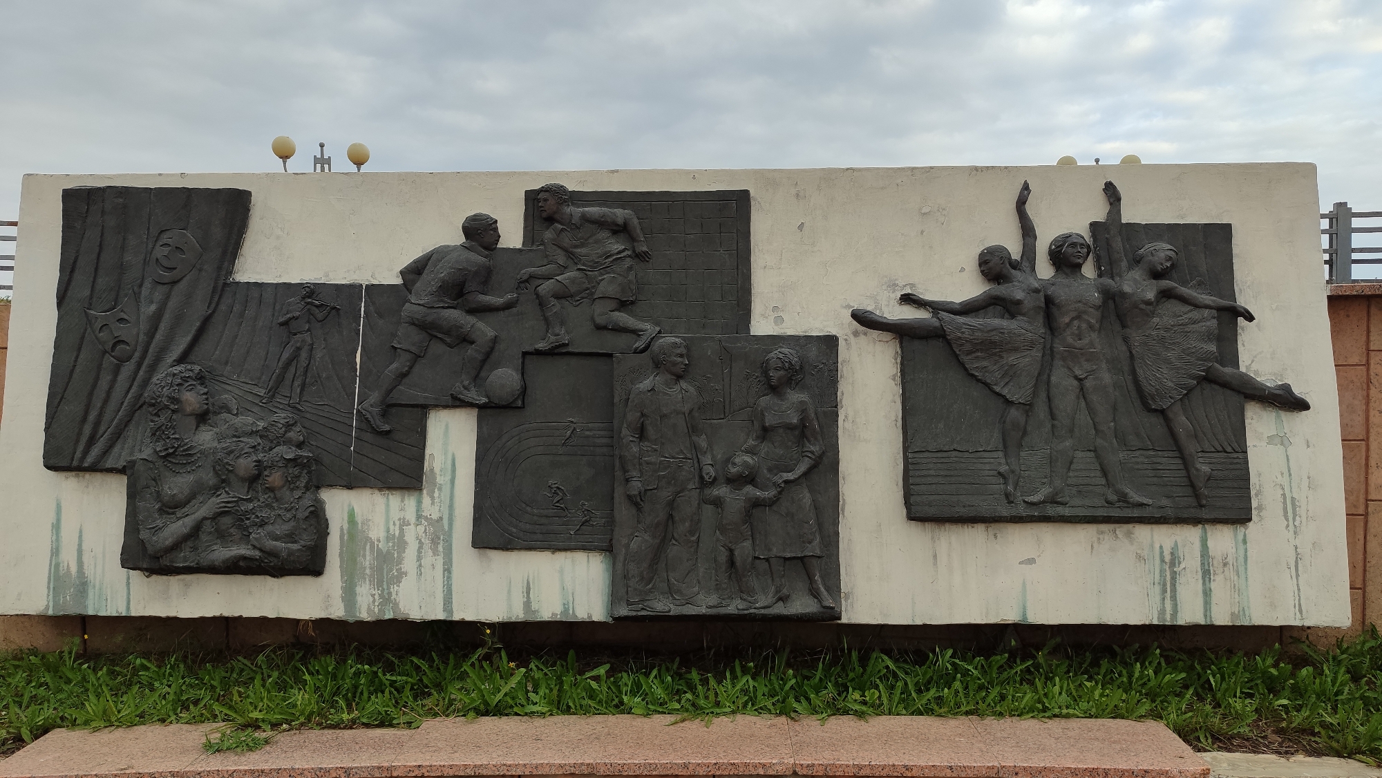 This is a photograph of a large, relief sculpture on a white wall, located in Mirny, Russia. The sculpture is divided into several panels, each depicting a different scene. From left to right, the panels show: a theatrical mask and curtains, a violin player and audience members, two men playing soccer, a family of three, and three ballerinas. The sculpture is made of dark-colored material and stands out against the white background. In the background, the sky is overcast with clouds. In front of the sculpture, there is a patch of green grass. The wall itself appears to be made of concrete and shows some signs of weathering. There are some lights mounted on poles above the sculpture. The sculpture is a public art installation, likely intended to represent various aspects of culture and life.