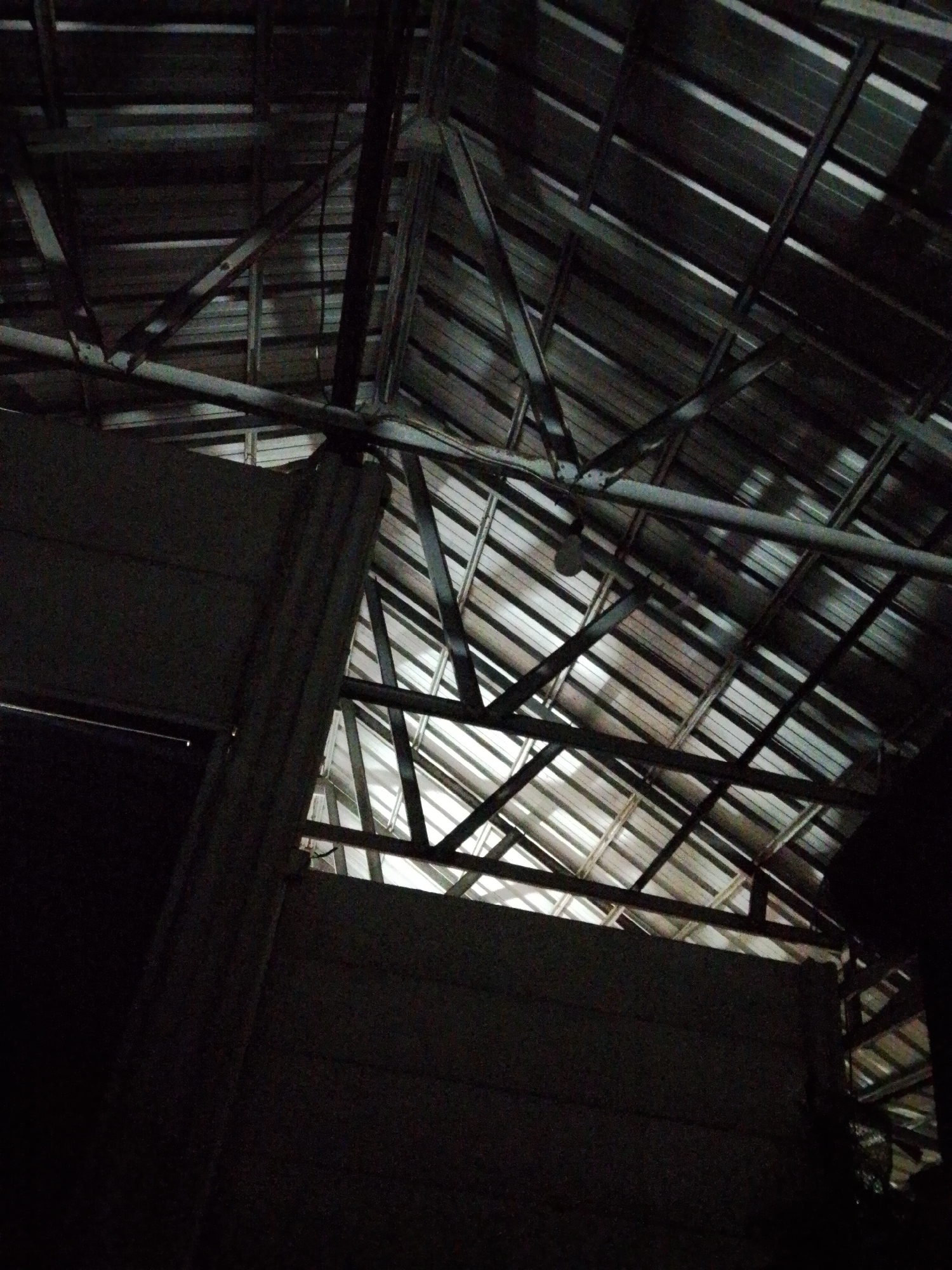 The image shows a low-angle view of the interior of a building or structure in Bukit Ketok, Indonesia. The scene is dimly lit, with the main light source appearing to be daylight filtering through gaps in the corrugated metal roof and structural supports. The roof is constructed of metal sheets with a grid of metal beams and supports underneath. The walls appear to be made of dark, possibly concrete or similar material. The scene gives the impression of an unfinished or industrial-style space. There are no people visible.