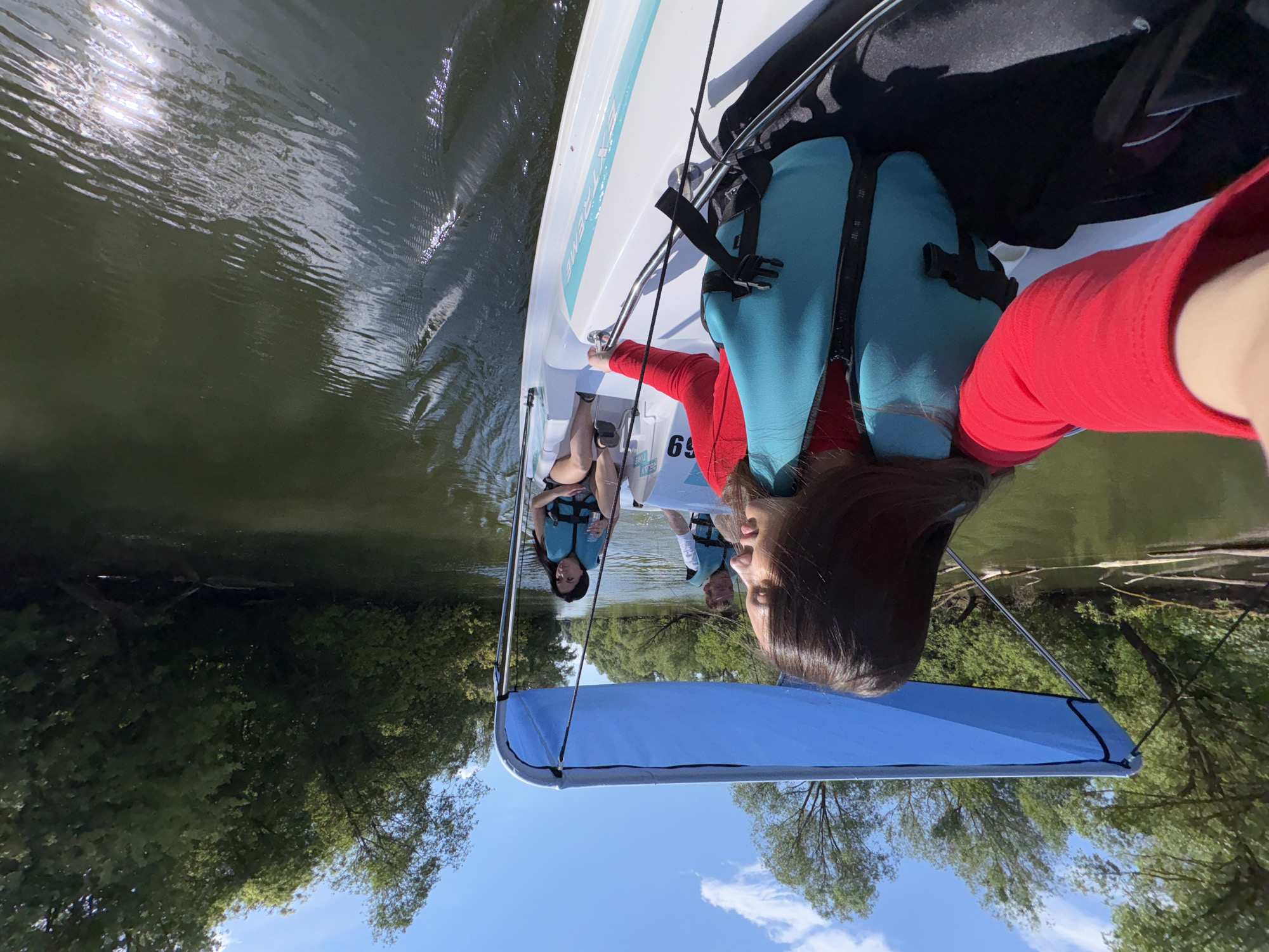 Four people are in a small boat on a river surrounded by trees under a blue sky with scattered clouds. The boat is white with a blue canopy. Two women and one man are wearing life jackets. The woman in the foreground is wearing a red shirt and is taking a selfie. The boat appears to be a pedal boat. The scene is likely a recreational area, possibly on the Volga River near Kazan, Russia.