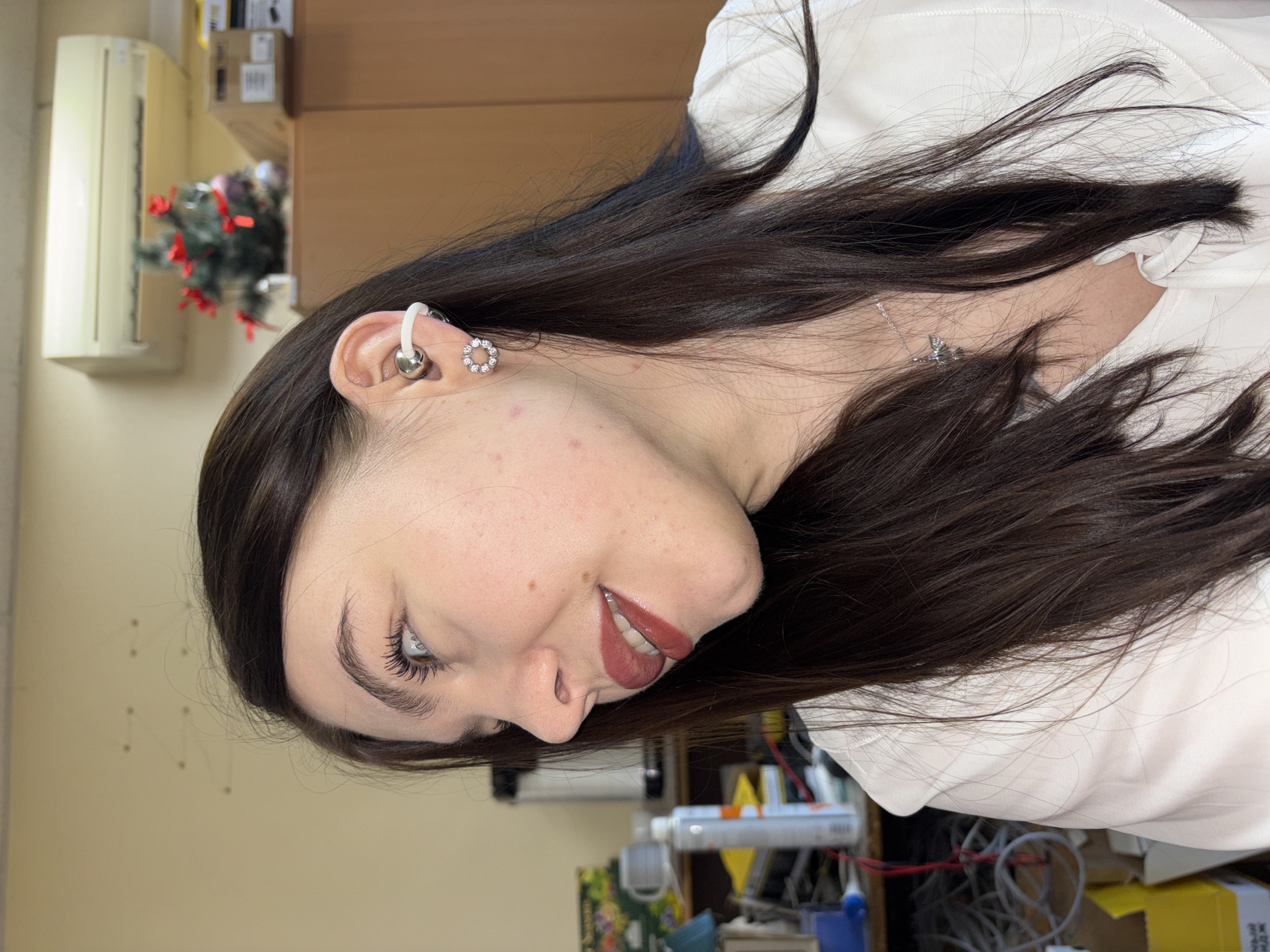 A young woman is seen in profile, likely indoors given the presence of office equipment and a window AC unit. She has dark hair and fair skin with some minor blemishes. She wears a white blouse, and several earrings in her left ear including a white cartilage earring, a silver bead stud, and a small decorative stud. A silver necklace is also visible. The background features a cluttered office scene with a wooden cabinet, a Christmas decoration, and various other items. The image may have been taken in Perm, Russia, based on contextual information.