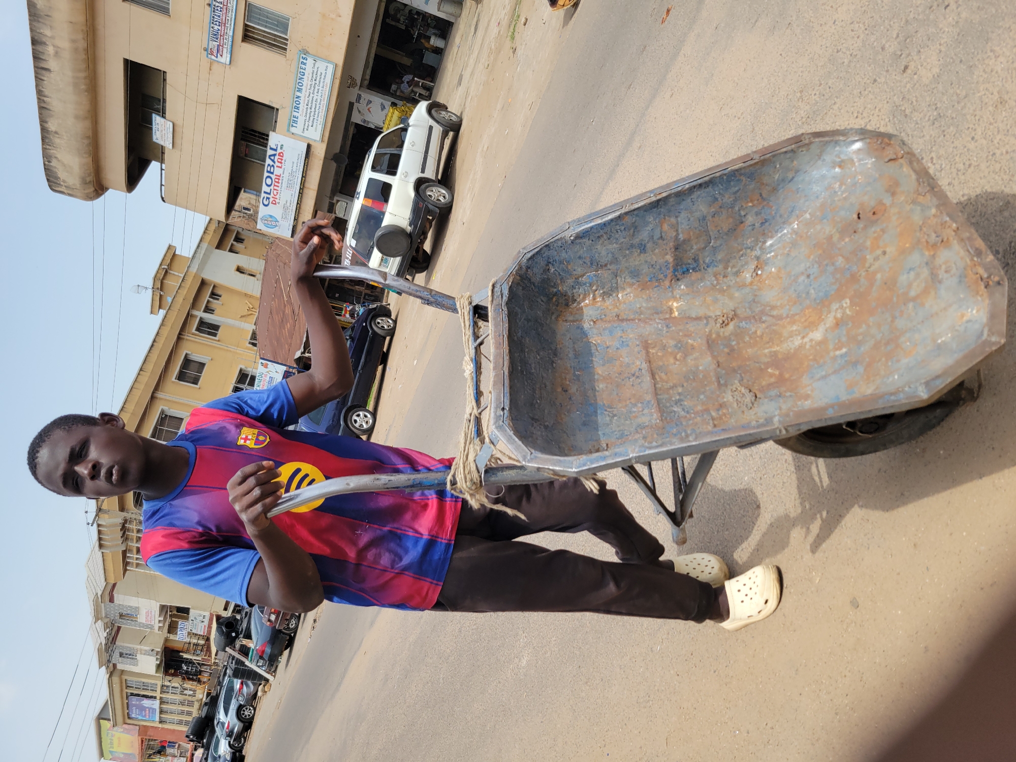 A young man in Jos, Nigeria, pulls a rusty, empty wheelbarrow on a paved road. He wears a Barcelona soccer jersey, black pants, and off-white Crocs. The wheelbarrow is blue with rust. The background shows buildings with signs for businesses including "GLOBAL DIGITAL LAB," "THE IRON MONGERS," and "VANAC ESTATES." Cars are parked along the street. The sky is overcast.
