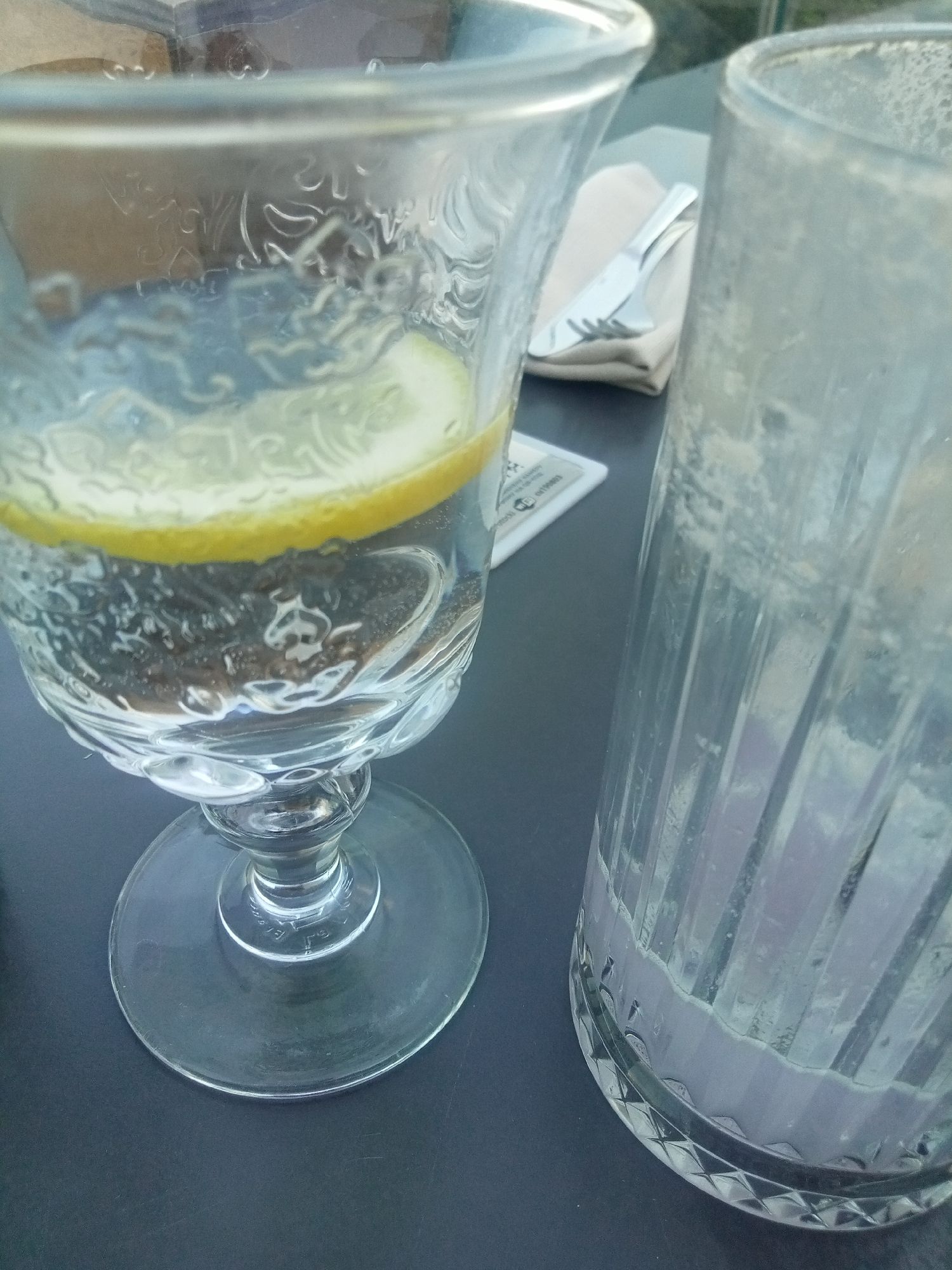An outdoor setting at ground level is captured, with the focus on two glasses on a dark surface, likely a table. The glass on the left is a stemmed wine glass filled with clear liquid and a slice of lemon. The glass on the right is a tall, fluted tumbler that appears to be empty or contains a small amount of liquid at the bottom. A folded napkin with cutlery is visible between the two glasses. The lighting suggests it is daytime, possibly late afternoon or early evening, with soft shadows and diffuse light. The background is blurred, but appears to be an outdoor environment with some foliage or structures. The overall impression is of a dining or drinking setup in an outdoor cafe or restaurant.