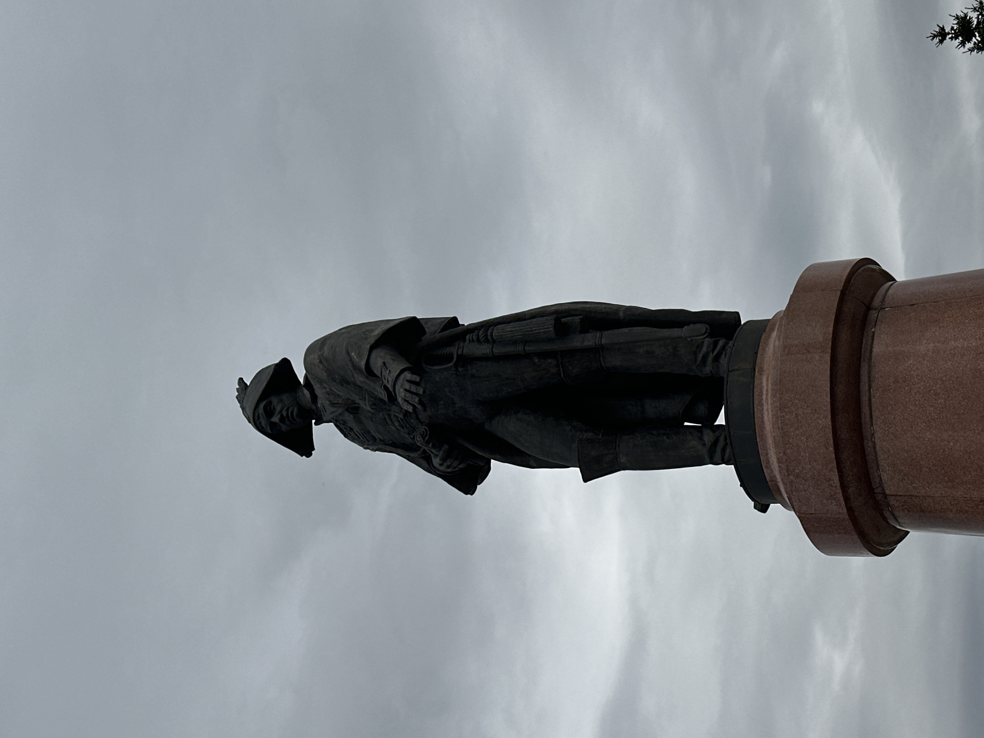 The image shows a statue of Vasily Surikov in Krasnoyarsk, Russia. The bronze statue stands atop a red stone pillar against a gray sky. Surikov is depicted wearing a hat, coat, and boots, and appears to be looking slightly downward. The statue is a prominent landmark in Krasnoyarsk.
