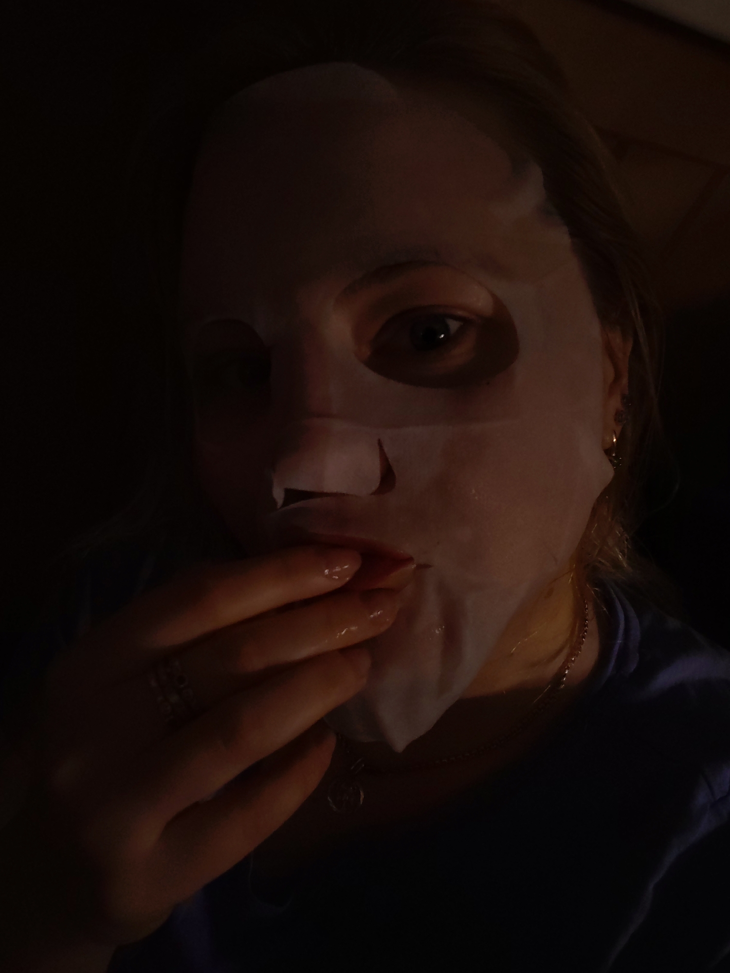A person is shown in a low-light setting wearing a sheet mask on their face. The mask covers most of the face, with holes cut out for the eyes. A hand, adorned with a ring, is near the mask, and a small amount of the person's neck is visible. The background is dark and indistinct. The location in Izhevsk, Russia cannot be identified from the given information.