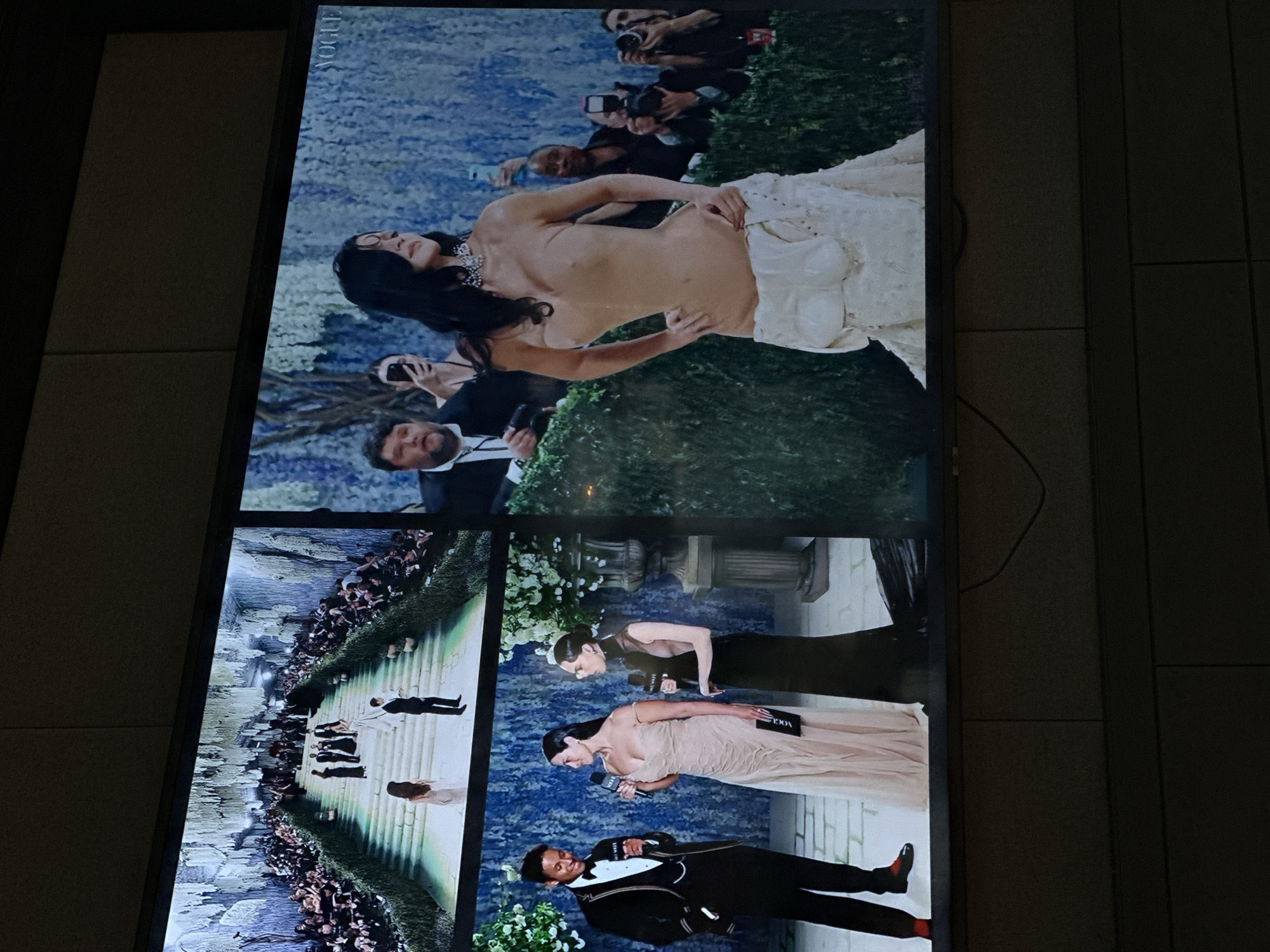 The image shows a television screen displaying Vogue magazine's coverage of the 2023 Met Gala in New York City, United States. The top portion of the screen shows a celebrity posing topless on the red carpet, while being photographed by members of the media. Other images show celebrities being interviewed, and a general scene of the red carpet arrivals. The television is located in a room with tiled walls.