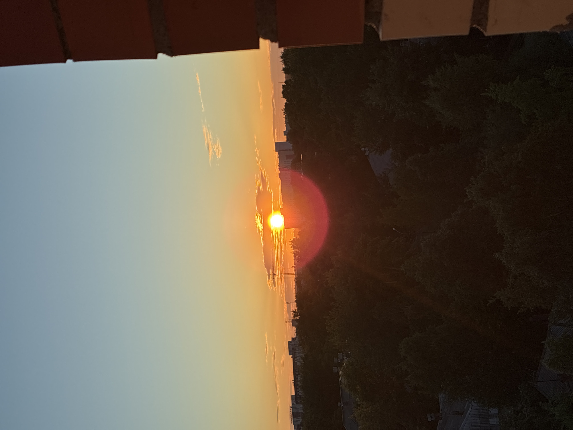 An outdoor, eye-level, vertical shot captures a sunset over a city. The sky transitions from a light blue at the top to a warm orange as it nears the horizon. A bright sun, partially obscured by clouds, sits just above a dense line of dark green trees that stretches across the middle of the frame. In the distance, silhouetted buildings and a construction crane are visible. A bright red lens flare emanates from the sun. The foreground features a brick structure on the right side of the image.