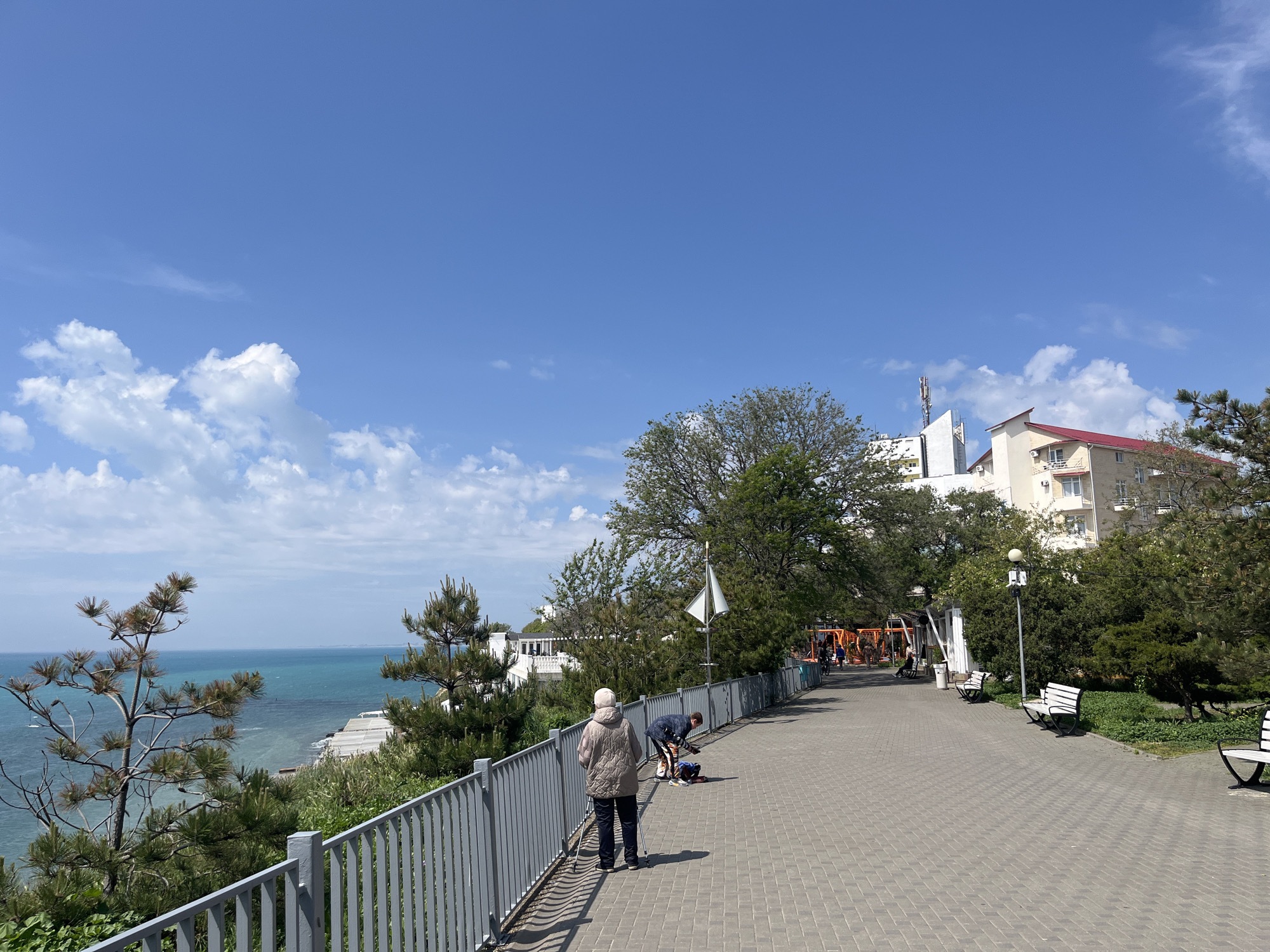 The image shows a paved walkway in Anapa, Russia, overlooking the sea. The walkway is lined with a grey metal fence, trees, and benches. An elderly woman with hiking poles is standing on the walkway, looking out at the sea. Nearby, a man is bent over, possibly attending to something on the ground. The sea is a turquoise color and there are white cliffs in the distance. The sky is blue with some white clouds. Buildings are visible behind the trees.