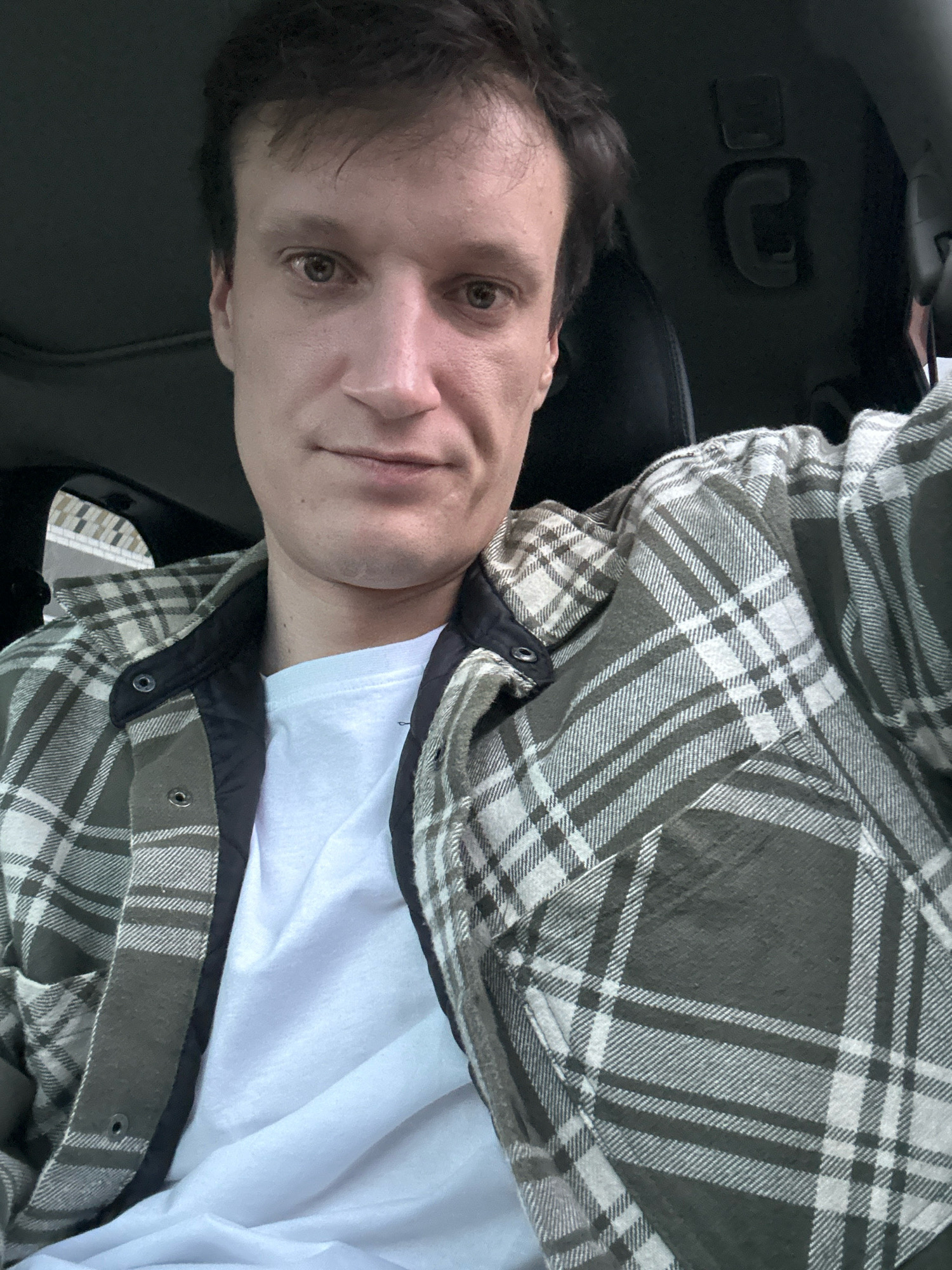 The image shows a close-up, selfie-style shot of a person sitting in the driver's seat of a vehicle. The person has brown hair and light skin, and is wearing a white t-shirt underneath a green and white plaid shirt jacket. The background is dark, suggesting the interior of the car, with some visible details of the car's roof and a handle. The lighting is moderate, with some shadows on the person's face. There is no visible indication of the location being Shatava, Ukraine.