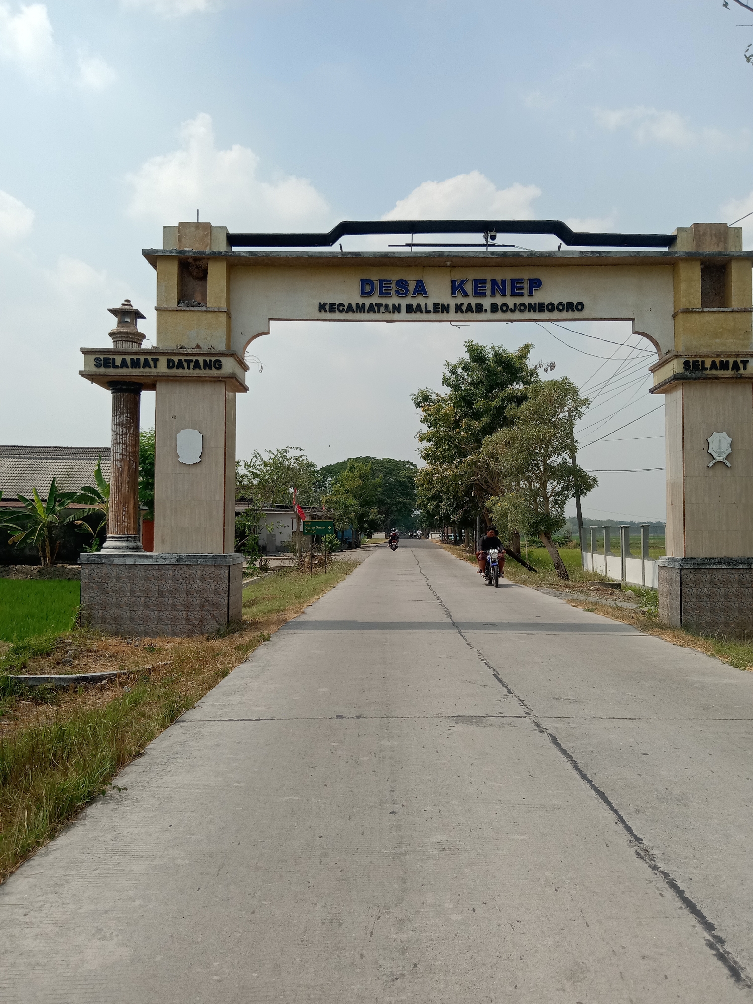 The image captures an archway marking the entrance to "DESA KENE P", indicating a village. The text below reads "KECAMATAN BALEN KAB. BOJONEGORO", specifying the sub-district and regency in Indonesia. On the left pillar, "SELAMAT DATANG" is written, meaning "Welcome". On the right pillar, "SELAMAT" is also visible, likely the start of "Selamat Jalan" or "Selamat Datang" repeated. A paved road leads through the archway, with a few motorcycles visible in the distance. The environment appears rural, with green fields and trees lining the road. The sky is partly cloudy. The archway is made of concrete with stone accents at the base of the pillars. There is a small, decorative structure on top of the left pillar and a crest on the front of both pillars. The scene is set during the daytime, under natural light. The exact location within the city of Mayangkawis, Indonesia is not specified in the image itself, but the provided text indicates the administrative region.