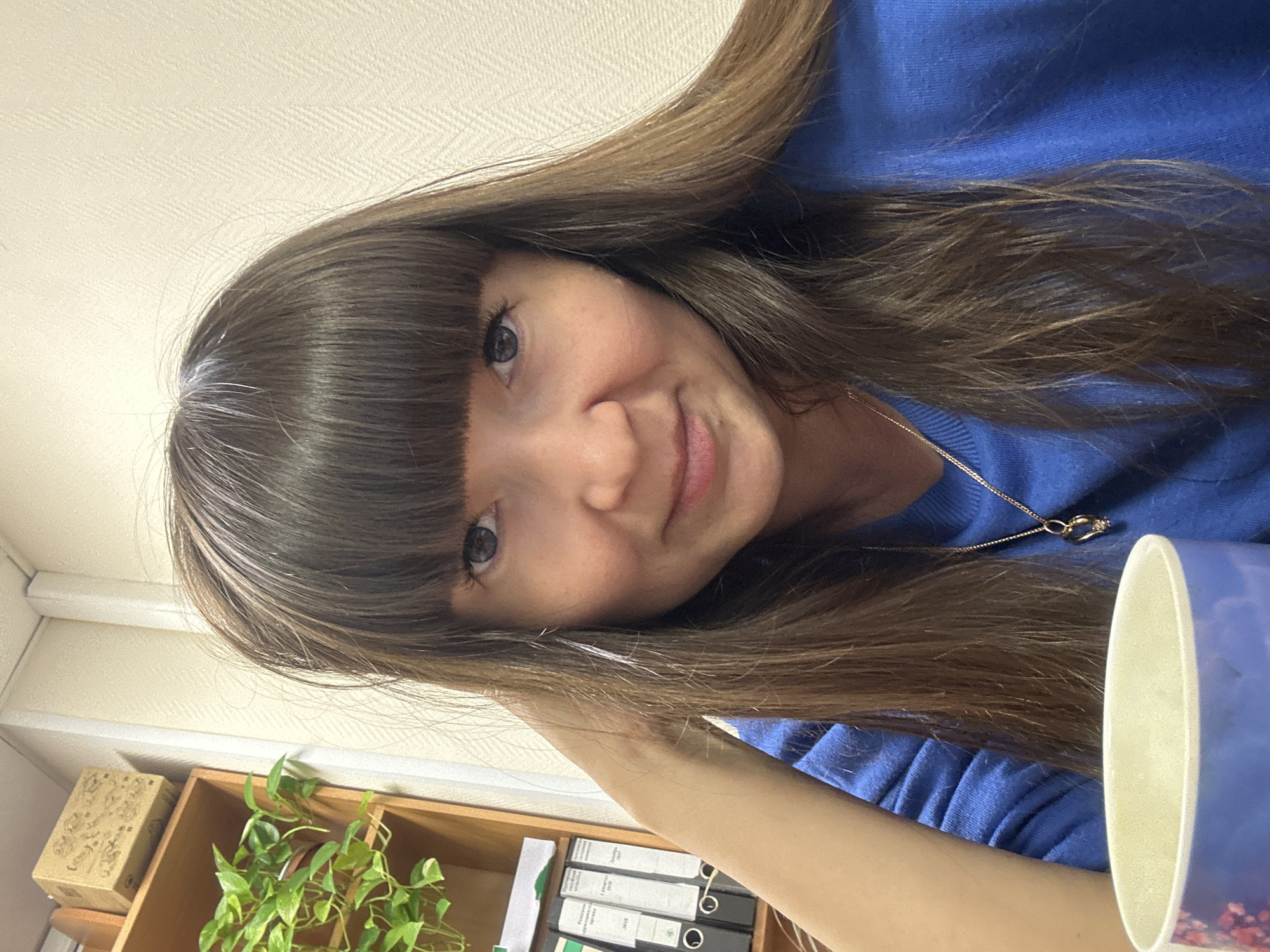 A selfie of a young woman with long brown hair and bangs, wearing a blue sweater, is oriented at an angle, with the top of her head towards the upper left and her right shoulder towards the lower right. A gold necklace with a small pendant is visible. A cup with a white interior and blue exterior is in the lower right. Behind her and to the left is a light-colored wall and a shelf unit containing a potted plant, some boxes, and ring binder files. I am unable to tell you that this image was taken in the city of Chaikovsky, Russia.
