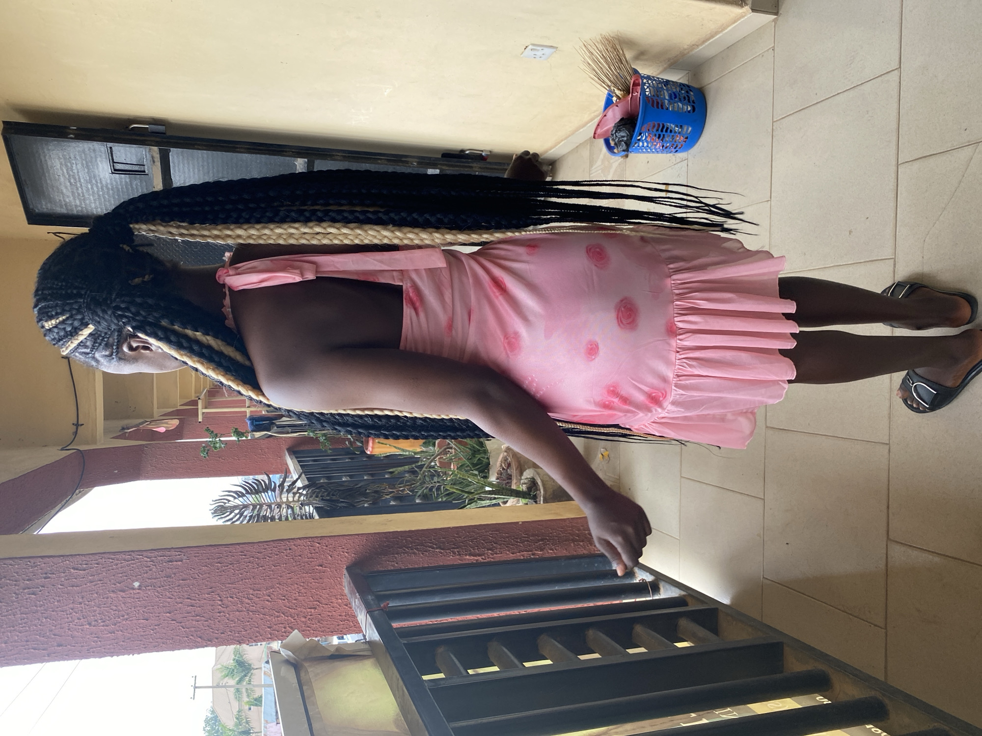 A full-length portrait, currently displayed on its side, features a dark-skinned individual standing outdoors or in a semi-outdoor residential space in Jos, Nigeria, under bright daylight. The person, likely a woman, has long, voluminous box braids with black and light blonde/gold sections. She is wearing a sleeveless, light pink dress with shoulder ties and a ruffled hem, adorned with subtle circular, rose-like patterns. Her feet are clad in dark sandals with a silver accent.
The setting includes a light-colored tiled floor and walls painted in light yellow/beige and a textured red-brown. In the background, to the upper right (when viewed upright), a blue plastic basket containing a broom, a pink dustpan, and a black plastic bag rests against the wall. A white power outlet is visible on the upper wall. To the upper left, a small, dark-framed window or vent can be seen. In the foreground, spanning the lower half of the image, is a dark metal railing, possibly part of a balcony or staircase. The person's right hand rests gently on this railing. Beyond the railing, glimpses of green plants, clear sky, and other buildings indicate an outdoor environment. The bright natural lighting suggests it is daytime with fair weather. No explicit activity other than standing is taking place, and no discernible text is visible in the image.