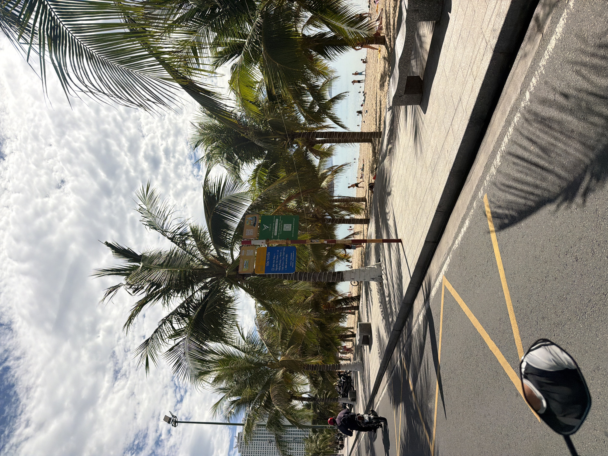The image depicts a vibrant coastal scene in Nha Trang Ward, Vietnam, captured from the perspective of someone on a motorcycle. The viewer is looking along a palm-lined boulevard with a sandy beach and the ocean on the right, and a paved road on the left.
Numerous tall palm trees with lush green fronds dominate the center of the frame, casting long, distinct shadows across the sidewalk and the road, indicating bright sunshine. The sky above is partly cloudy with scattered white cumulus clouds.
On the beach, several people are visible, some standing in the shallow water of the ocean, others strolling on the sand, enjoying the sunny day. The ocean appears calm and light blue. A concrete sidewalk separates the palm trees and beach from the asphalt road.
On the road, at least two motorcycles are present, with one rider wearing a red helmet partially visible on the left side of the frame. The road features yellow painted lines, possibly indicating parking or traffic lanes. In the distance, towering urban buildings are visible, suggesting a developed city backdrop.
Two informational signs are attached to palm tree trunks near the road. The OCR text for these signs reads: "TÊN TUYẾN" (Route Name) followed by a list of locations or street names: "Bắc Hàn Ông," "Nguyễn Tất Thành Lê," "Hồng Phong Tersin," and "Trần Phú - Tôn Thất Tùng." These are likely local street or route markers. The overall atmosphere is bright and lively, characteristic of a popular beach destination.