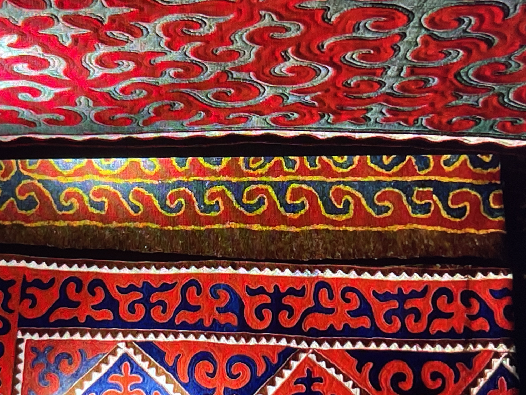 The image displays a close-up view of intricately patterned textiles, likely traditional Kyrgyz fabrics. On the left, a section of a textile features a repeating geometric pattern in red and blue, with a white serrated border. Adjacent to this is a vertical band of fabric with a red background and a yellow and blue swirling motif. To the right, a third textile is visible, predominantly red with a textured, greyish-blue background, also displaying a swirling, organic pattern. The lighting suggests the textiles are illuminated from the front, highlighting the textures and vibrant colors. The setting is likely an indoor display or a market stall in Bishkek, Kyrgyzstan, where such handicrafts are commonly sold. There are no people or discernable actions visible in the frame.