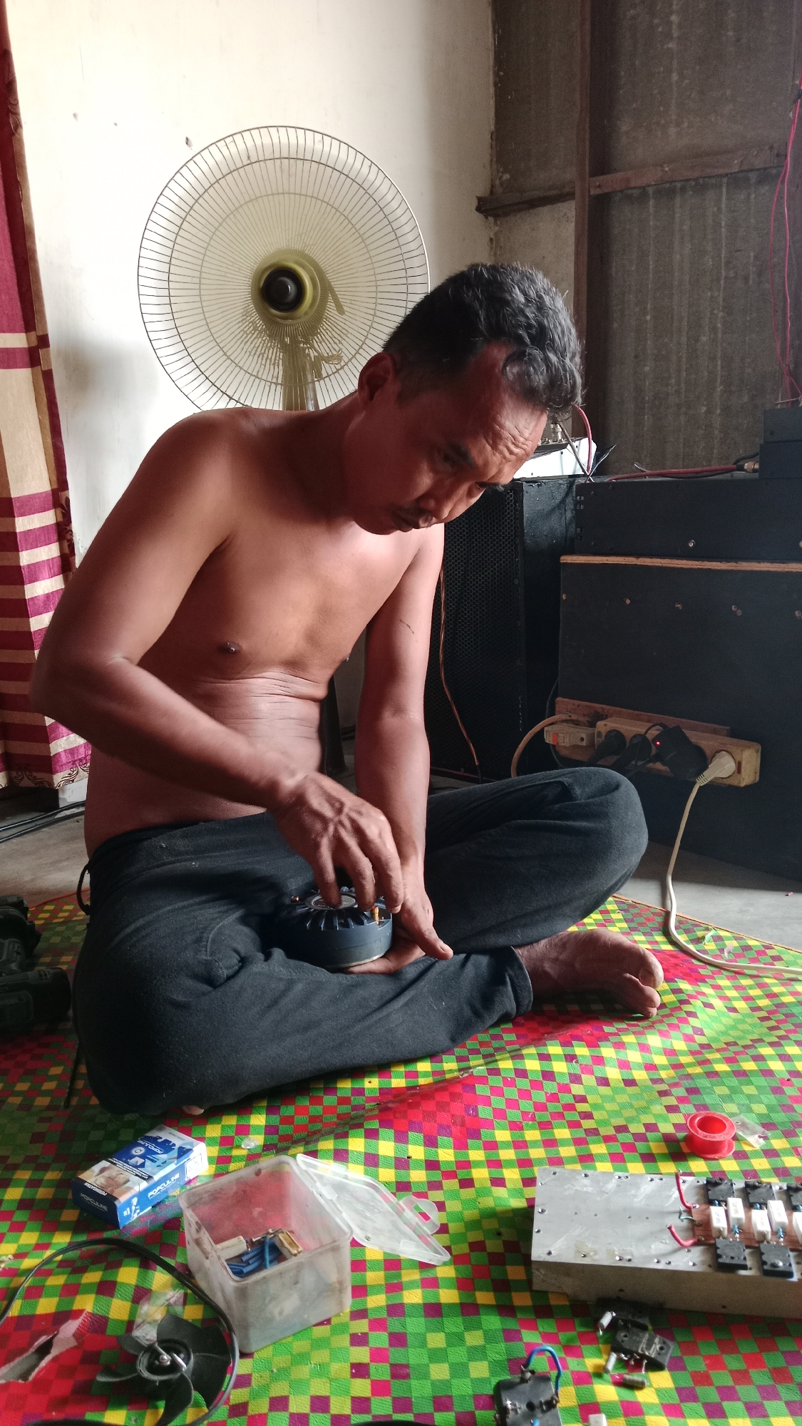 In a dimly lit interior, likely in Karang Bunga, Indonesia, a shirtless man is seated cross-legged on a colorful checkered mat. He is focused on a small, dark, circular electronic component held in his left hand, appearing to be working on it with his right hand. The man wears dark pants.

The environment suggests a makeshift workspace. To the left, a white oscillating fan is positioned against a light-colored wall, partially obscured by a striped curtain. Behind the man, two large black speaker cabinets are visible. One speaker cabinet has a power strip attached to its side, with several plugs inserted. Electrical wires are also apparent.

Scattered on the mat around the man are various objects: a pack of cigarettes, a clear plastic container with electronic components, a red spool of tape, and a metal box with multiple electrical terminals and wires. The floor beneath the mat appears to be concrete. The overall impression is one of repair or assembly of electronic equipment in a domestic or workshop setting.