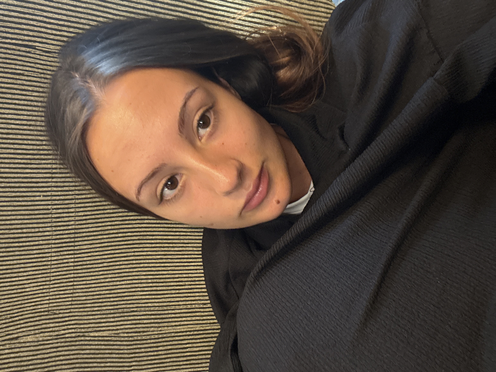 In Skala Marion, Greece, a young woman with long, dark hair is captured in a close-up, indoor portrait. She is positioned on a couch with a vertically striped fabric in shades of dark green and beige behind her. The woman is wearing a black garment that covers her chest and shoulders. Her gaze is directed slightly off-camera, with one eye more visible than the other due to the angle. Her skin tone appears warm, and she has a mole visible on her lower lip. The lighting suggests an indoor setting, possibly a home, with natural light illuminating her face.