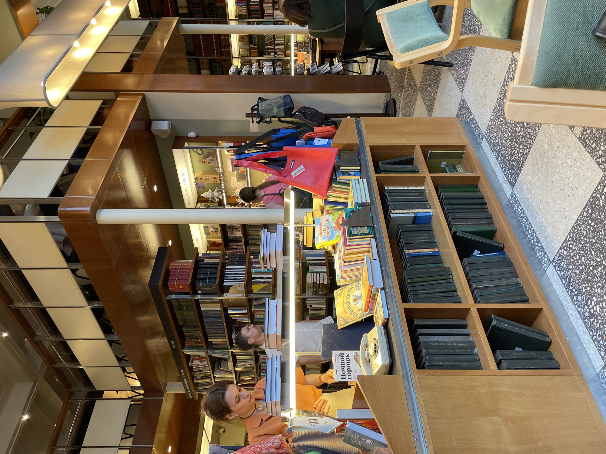 This image depicts the interior of a multi-level bookstore or library located in Saint Petersburg, Russia. The scene is characterized by extensive wooden bookshelves filled with books, spanning from the ground level to an upper mezzanine. The floor features a checkerboard pattern composed of alternating light speckled and dark speckled terrazzo tiles.
In the foreground, a young woman with light brown hair, wearing an orange short-sleeved top and a dark choker necklace, stands behind a light wooden display counter. She is observed looking down at several stacks of books on the counter, with her left hand resting on one stack. Prominently displayed book covers include "Ночной горшок" (Night Pot) and "Утренний город" (Morning City). A white vintage-style rotary telephone is placed on the counter. Below the counter, shelves contain uniformly dark-covered books, some with Cyrillic titles such as "СМЫСЛ • ДНЕВНИК • ПИСЬМА ТУ СТЕМЫ." Two light wooden chairs with light green fabric upholstery are partially visible in the bottom right corner.
In the mid-ground, a man with short hair and sunglasses on his head, wearing a white short-sleeved shirt, stands behind the woman in orange, observing something to his right. Further back, a woman in a pink top is partially visible near a display area. To the right, a metal stand holds several tote bags in red, blue, green, and black. Another woman, wearing a dark green top, sits at a table in the background right, her back partially turned towards the viewer.
Architectural elements include light-colored pillars supporting a wooden mezzanine level with wooden undersides. The mezzanine railing consists of a wooden top rail, metal bars, and white opaque panels. The interior is brightly illuminated by recessed spotlights on the ceiling and beneath the mezzanine, as well as a long rectangular light fixture with multiple lamps. Signs above various sections of the bookshelves are in Cyrillic, indicating categories such as "ПОЭЗИЯ" (Poetry), "КНИГИ ИЗ ИНОСТРАННЫХ ЯЗЫКОВ" (Books from Foreign Languages), "КАНЦЕЛЯРИЯ, ПОДАРКИ" (Stationery, Gifts), "ДЕТСКИЕ КНИГИ" (Children's Books), and "ТЕАТР" (Theatre). The "КАНЦЕЛЯРИЯ, ПОДАРКИ" display includes small wooden house models and a white gnome figurine. Some wall sections are painted a light sage green.