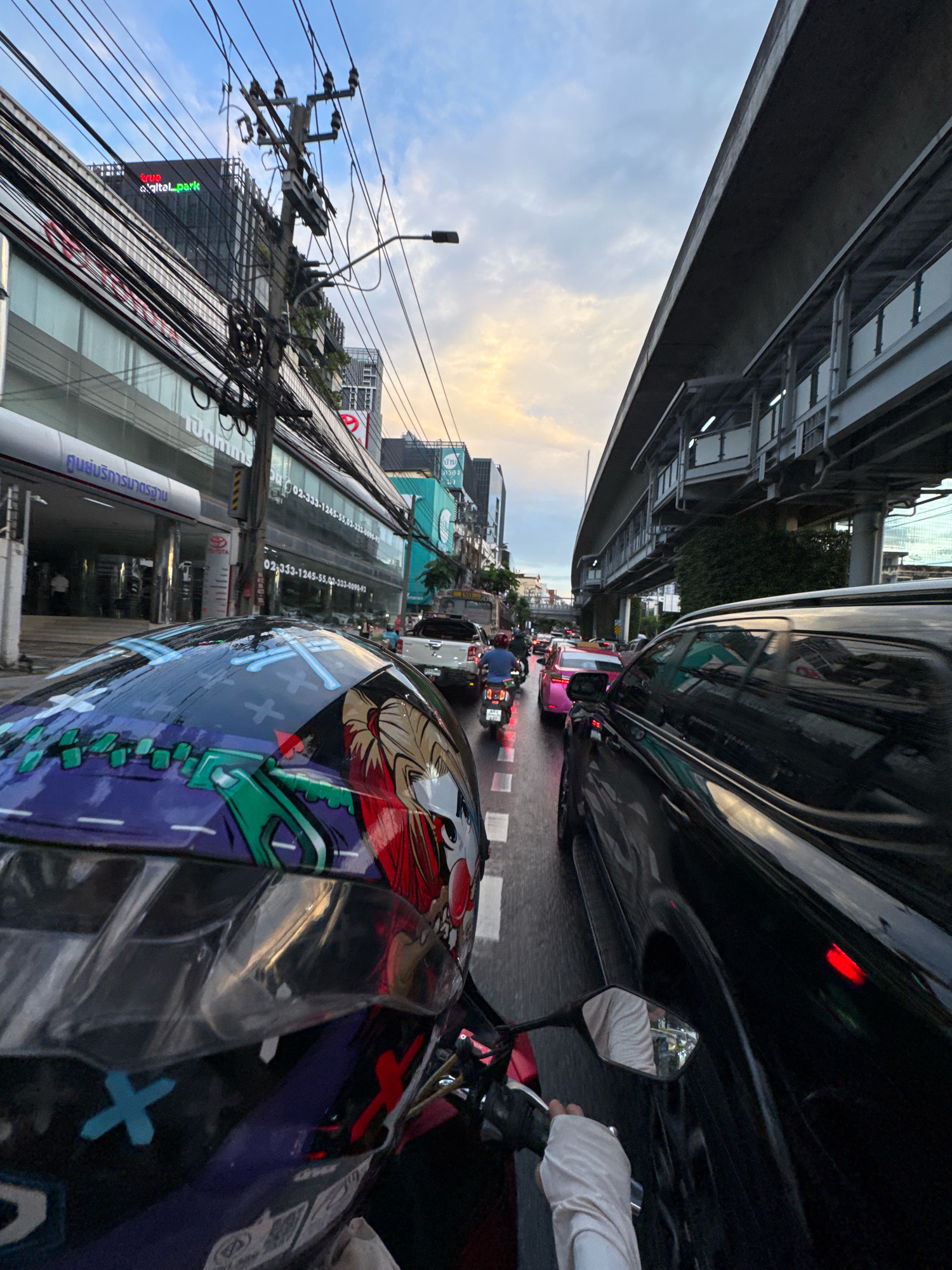 The image depicts a street scene in Bangkok, Thailand, during what appears to be rush hour. The perspective is from the viewpoint of a motorcycle rider, as indicated by the presence of a helmet in the foreground with a colorful anime-style design. The rider's gloved hand is visible on the handlebar.
The road is congested with vehicles, including cars, a white pickup truck, and several motorcycles. A pink car is also visible in the traffic. On the right side of the image, a large black SUV is prominently featured. Overhead, a modern elevated train or skywalk structure runs parallel to the road, with supporting pillars and tracks visible.
The surrounding environment shows urban architecture. Buildings line the street, with commercial signage and advertisements. One building in the background displays the logo "true digital park" in illuminated green and red text. Power lines and utility poles are densely strung across the sky. The sky itself is partly cloudy with a soft, warm light suggesting either sunrise or sunset. The overall impression is one of a busy, dynamic city environment.