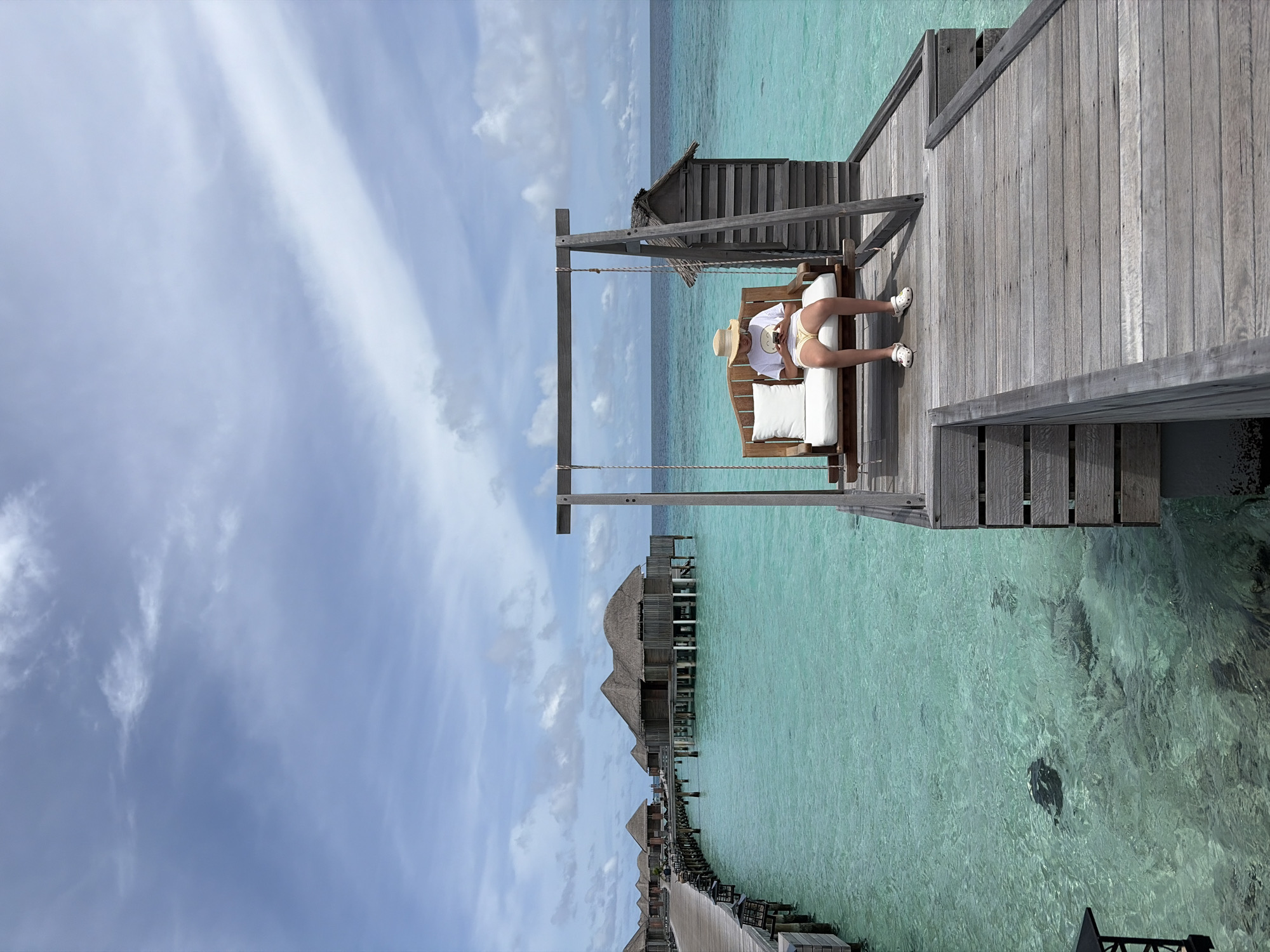 The image depicts a person sitting on a wooden swing on a pier in Halaveli, Maldives. The person, wearing a straw hat, white t-shirt, and yellow shorts, is looking down at a mobile phone. The swing is positioned over clear turquoise water, revealing the sandy bottom and some coral formations. In the background, overwater bungalows with thatched roofs stretch along the pier, characteristic of the resort's architecture. The sky is partly cloudy with a bright blue expanse. The wooden pier extends into the frame, leading the eye towards the bungalows.