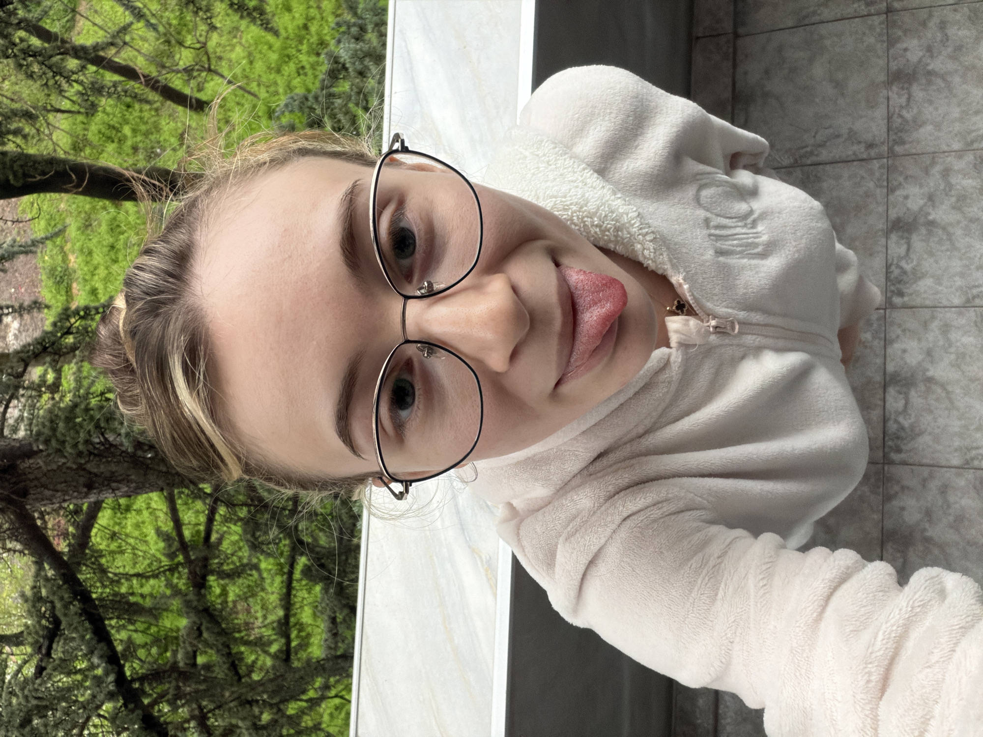 A young woman with blond hair and wearing black-rimmed glasses is looking directly at the camera and sticking out her tongue. She is wearing a white, fuzzy hooded sweatshirt. The background includes greenery, possibly trees, and a gray tiled wall or floor. The location cannot be determined.