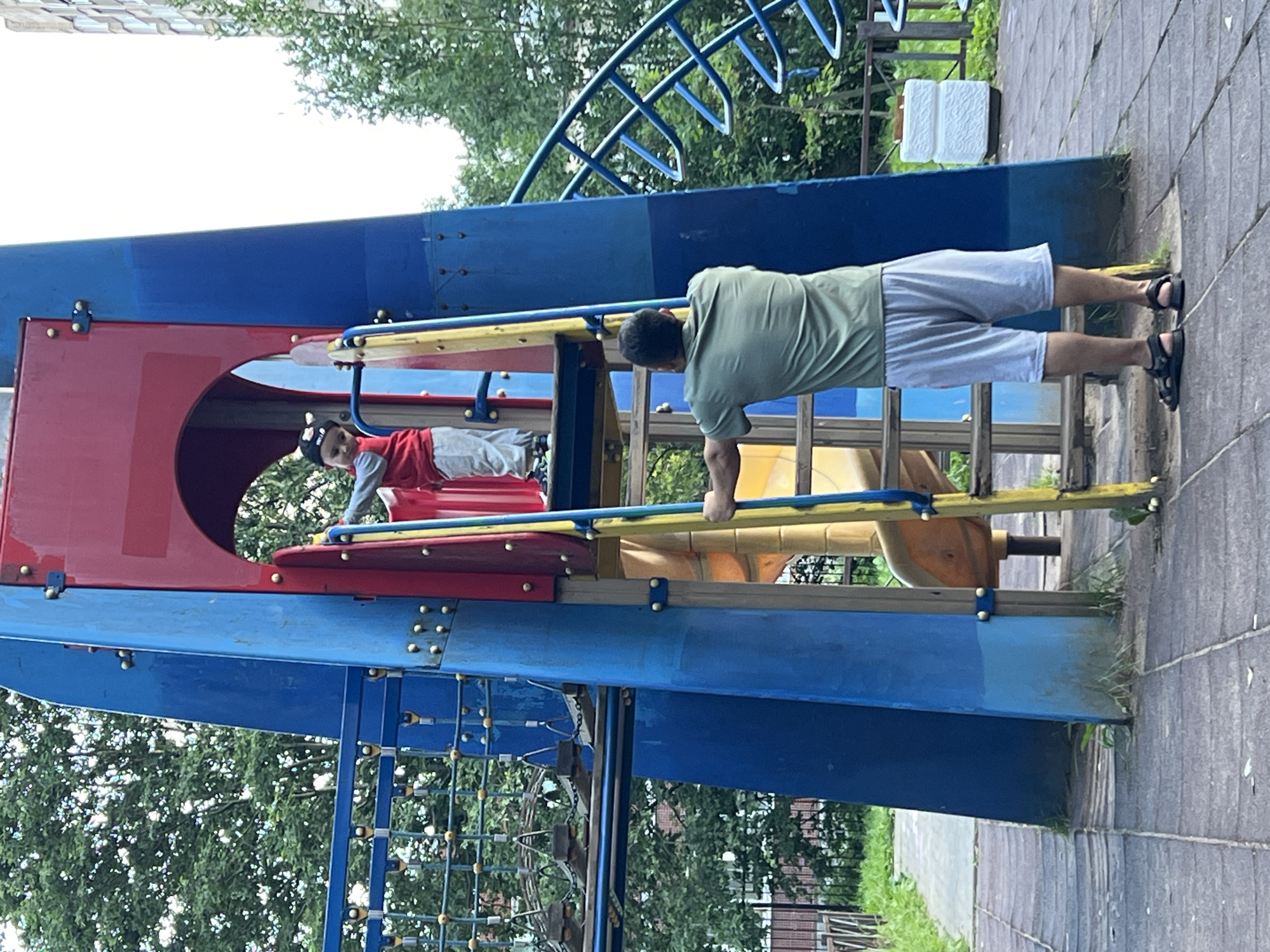 The image shows a playground scene in Sinyavino, Russia. A man in a green t-shirt and light gray shorts is performing a plank exercise on the playground structure. He is positioned with his hands on a yellow bar, his body extended parallel to the ground, and his feet resting on the ground. To his left, inside a red and blue section of the playground, a child wearing a red shirt and a black hat with white accents is visible. The child appears to be lying down.
The playground structure is predominantly blue and red, with yellow accents on the bars and ladders. Behind the structure, trees with green foliage are visible, suggesting it is daytime and likely a pleasant weather day, given the outdoor activity. The ground around the playground is paved with gray square tiles. A white, rectangular object, possibly a piece of playground equipment or a decorative element, is visible in the background on the paved surface. The overall scene depicts a recreational area with an adult engaging in exercise while a child is present. There is no visible text in the image.