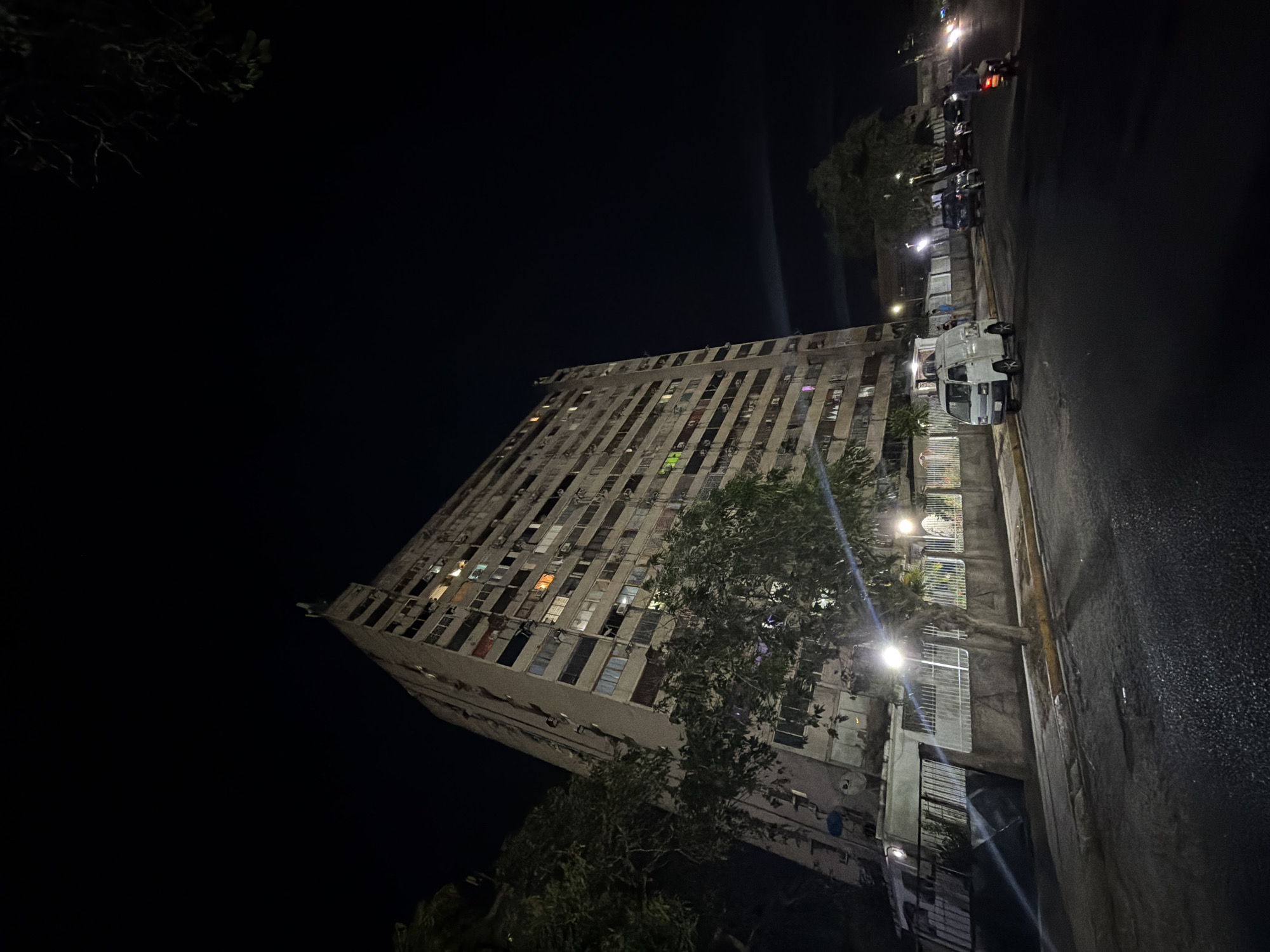 A nighttime exterior shot shows the Centro Civico building in Guayana City, Venezuela. The multi-story, weathered building is lit from within with lights in various windows. A few streetlights cast light on the building and street. A white truck is parked next to a sidewalk near the building's base. Trees are present, partially obscuring parts of the building. Other vehicles, including motorcycles and cars, are parked further down the street. The sky is dark.