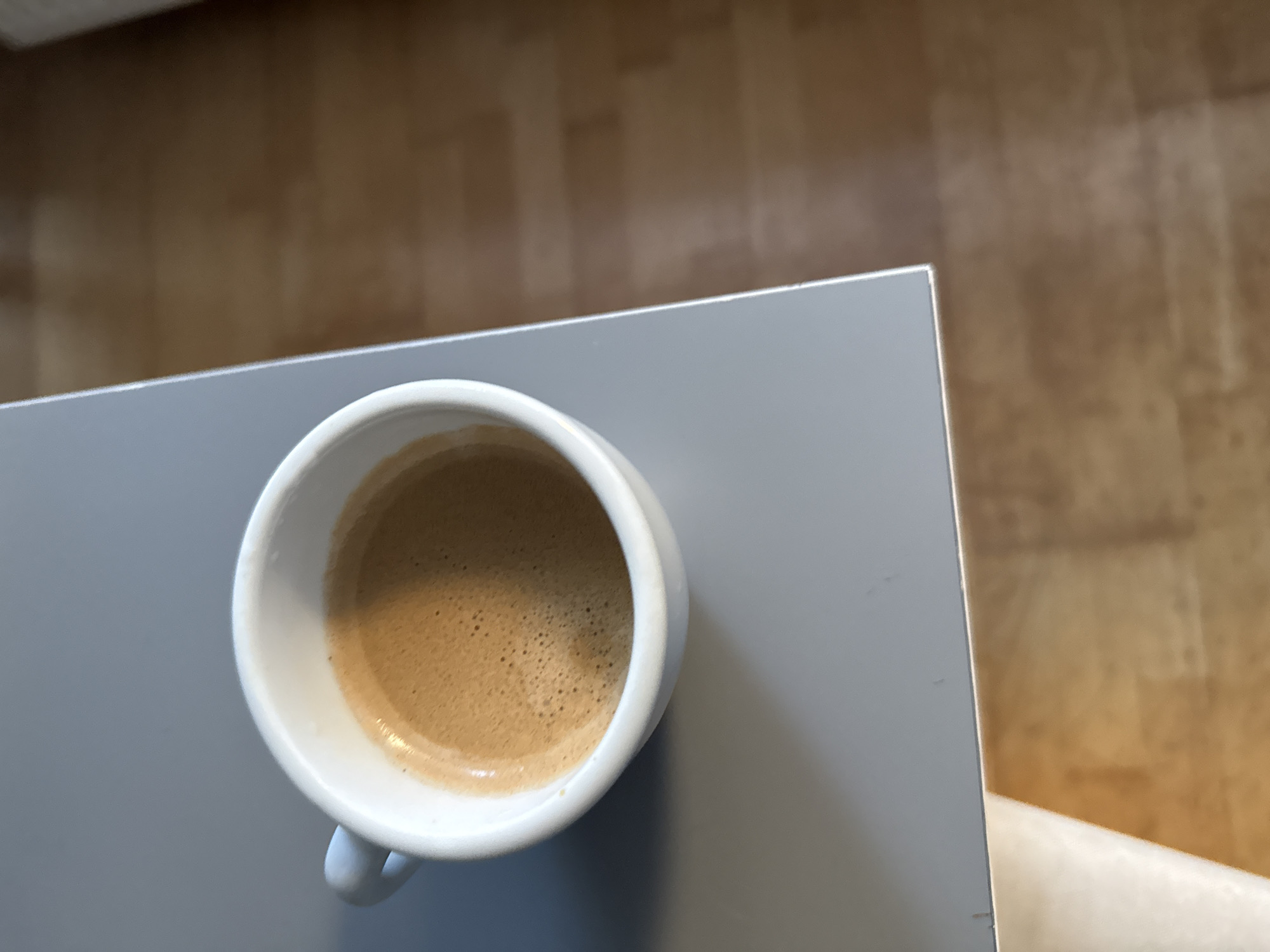 An espresso is in a white ceramic cup with a handle, resting on a gray surface. The espresso is dark brown with a layer of crema on top. The gray surface appears to be a table or countertop, and it is adjacent to a wooden floor. The lighting suggests it is daytime, with natural light coming from an unseen source. The image is taken from a top-down perspective. There is no visible information that allows for the determination of the city of Wrocław, Poland.