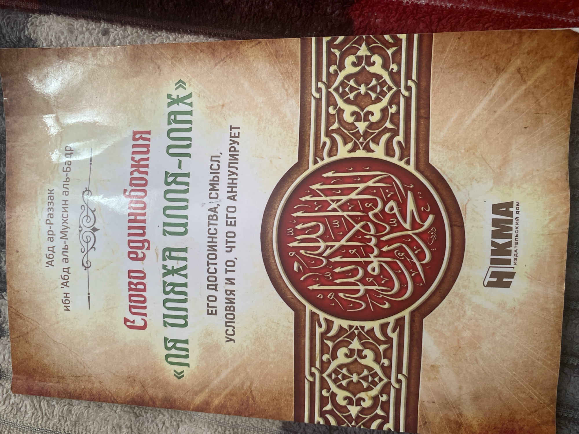 The image shows the cover of a book with a title written in Russian and Arabic calligraphy. The Russian title reads "СЛОВО ЕДИНОБОЖИЯ" which translates to "The Word of Monotheism". Below this, in green lettering, is what appears to be the transliteration of an Arabic phrase, "ЛЯ ИЛЯХА ИЛЛЯ-ЛЛАХ". Further text in Russian states, "ЕГО ДОСТОИНСТВА, СМЫСЛ, УСЛОВИЯ И ТО, ЧТО ЕГО АННУЛИРУЕТ", which translates to "Its Virtues, Meaning, Conditions, and What Nullifies It". The cover features ornate decorative elements, including Arabic script within a circular emblem and geometric patterns. The book cover is laid on a textured surface, possibly a blanket or fabric, in a muted color palette of beige, brown, and red. The bottom of the cover displays a logo with the word "HIКМА" and "ИЗДАТЕЛЬСКИЙ ДОМ" (Publishing House) in Russian. There is no indication of the city of Kizilyurt, Russia, or any other specific location in the image.