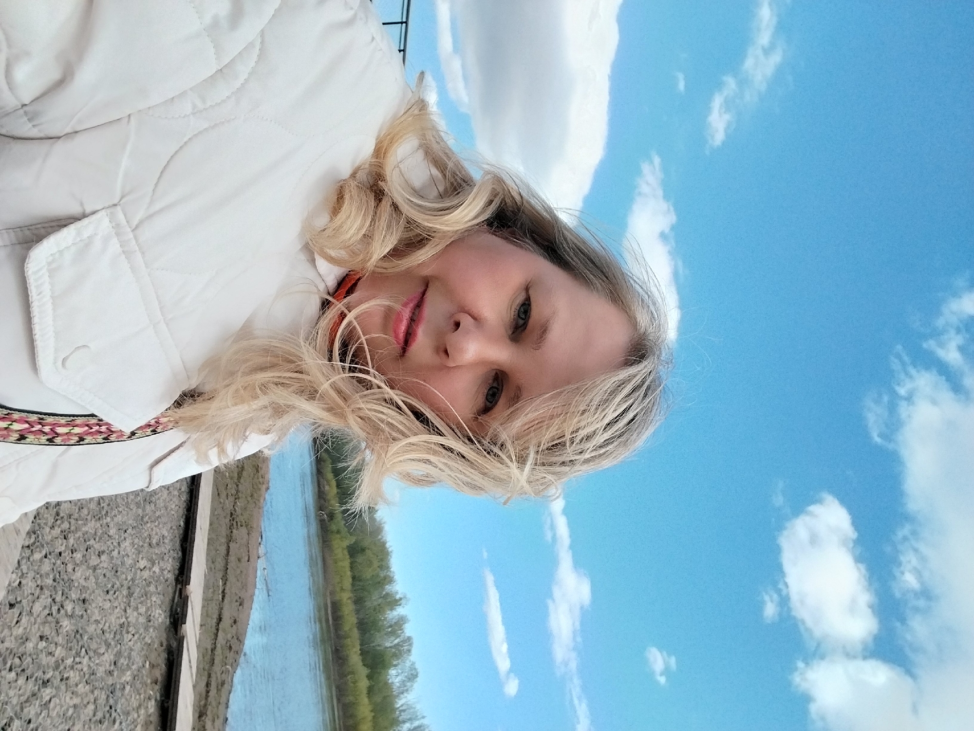 The image displays a bright outdoor selfie of a blonde woman, with the photograph itself rotated 90 degrees clockwise. The woman's head and upper body occupy the left side of the frame, with her blonde, wavy hair framing her face. She looks directly at the viewer with blue-gray eyes and a faint smile, her lips colored with pink lipstick. She is dressed in a light-colored, possibly white or off-white, quilted jacket, which features a decorative, colorful patterned band visible near the bottom left edge of the frame.
The background is vertically split. The upper right portion is dominated by a clear blue sky with scattered white cumulus clouds, indicating sunny daytime weather. Below the sky, a calm body of water, likely a river, stretches horizontally across the middle of the frame, with a line of green trees visible on its far bank. The bottom left segment of the image shows a dark gray, textured surface, appearing to be gravel or rocky ground on the near bank. The scene conveys a tranquil outdoor moment by a riverside, consistent with the provided location context of Orenburg, Russia. No visible text is present in the image.