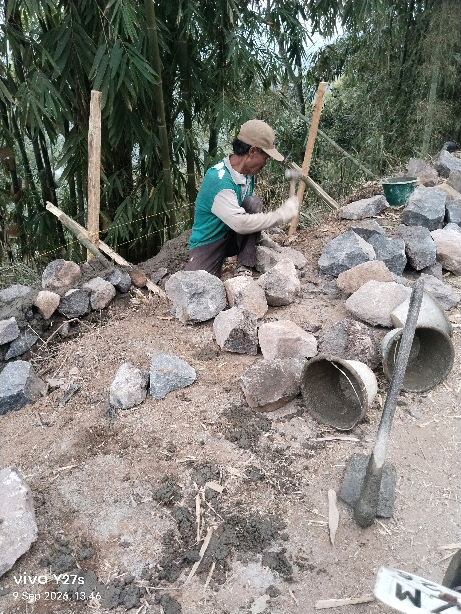 In Tamansari, Indonesia, a man is engaged in the construction of a rock wall. He is wearing a tan baseball cap, a green and white long-sleeved shirt, and dark pants. He is crouched down, using a hammer to strike a wooden stake that is positioned at an angle. The stake is partially embedded in the ground and secured with bamboo and twine. The man is surrounded by a collection of large, rough-hewn rocks of various gray and brown hues. Some of the rocks are already placed in the wall, held together by wet cement. Nearby, there are two buckets and a trowel, also covered in cement, suggesting ongoing construction work. The environment is outdoors, with a dense growth of bamboo trees in the background. The ground is composed of dirt and small debris. The image is dated September 9, 2026, at 13:46.