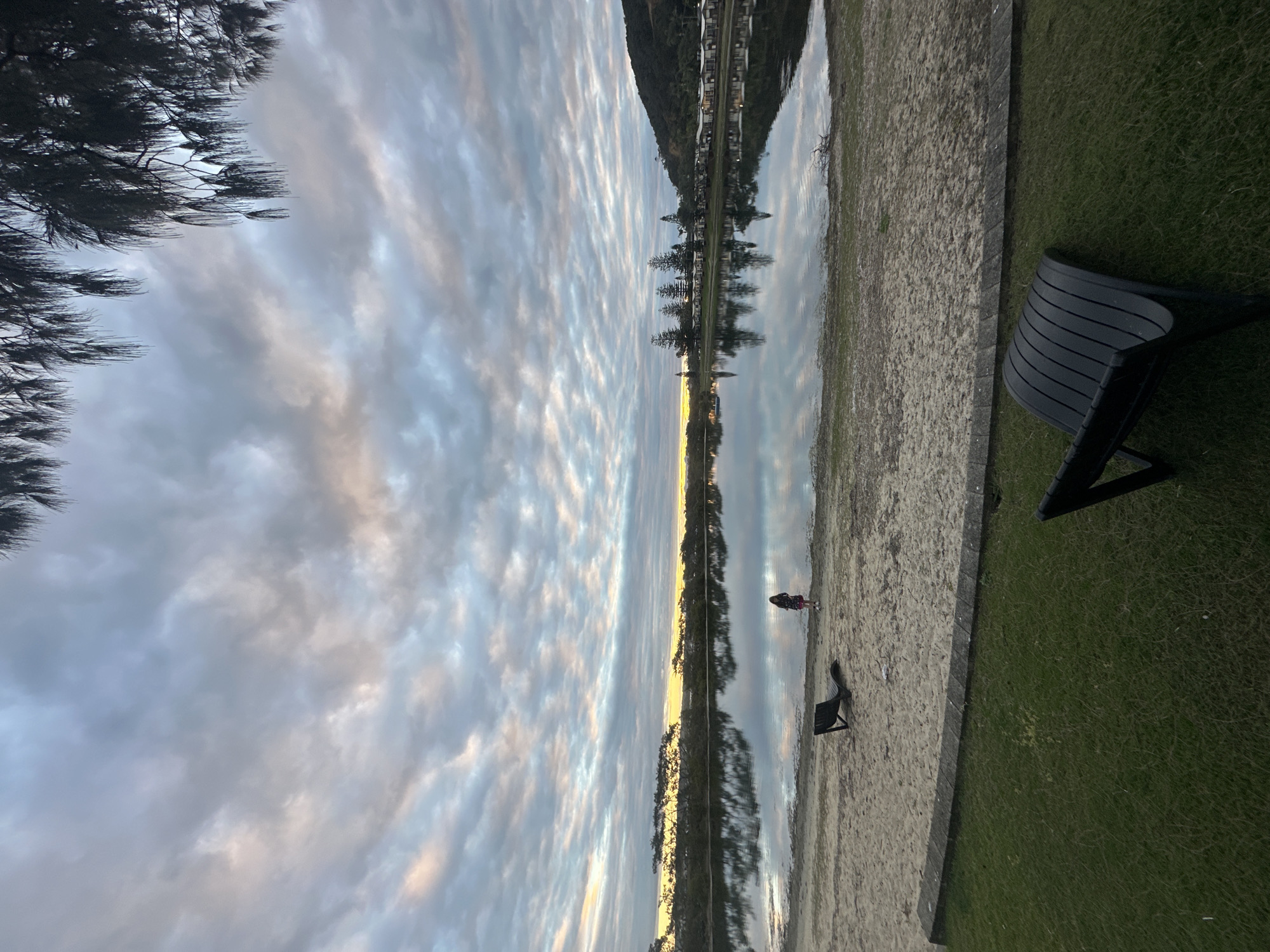 This is an image of the Nambucca River in Nambucca Heads, Australia, taken during twilight. The sky is filled with dramatic, textured clouds that are reflecting the muted colors of the setting or rising sun, with streaks of pink and orange visible amongst the grays and blues. The still water of the river acts as a mirror, reflecting the sky, the treeline on the opposite bank, and a row of buildings with peaked roofs. In the foreground, there is a strip of manicured green grass. Beyond the grass is a sandy area, and further back, a dark-colored lounge chair is positioned on the sand. A person, appearing to be a child, is standing near the water's edge on the sandy bank, facing away from the camera. Another dark lounge chair is visible in the foreground on the grass. The overall scene conveys a sense of tranquility and the soft, diffused light of twilight.