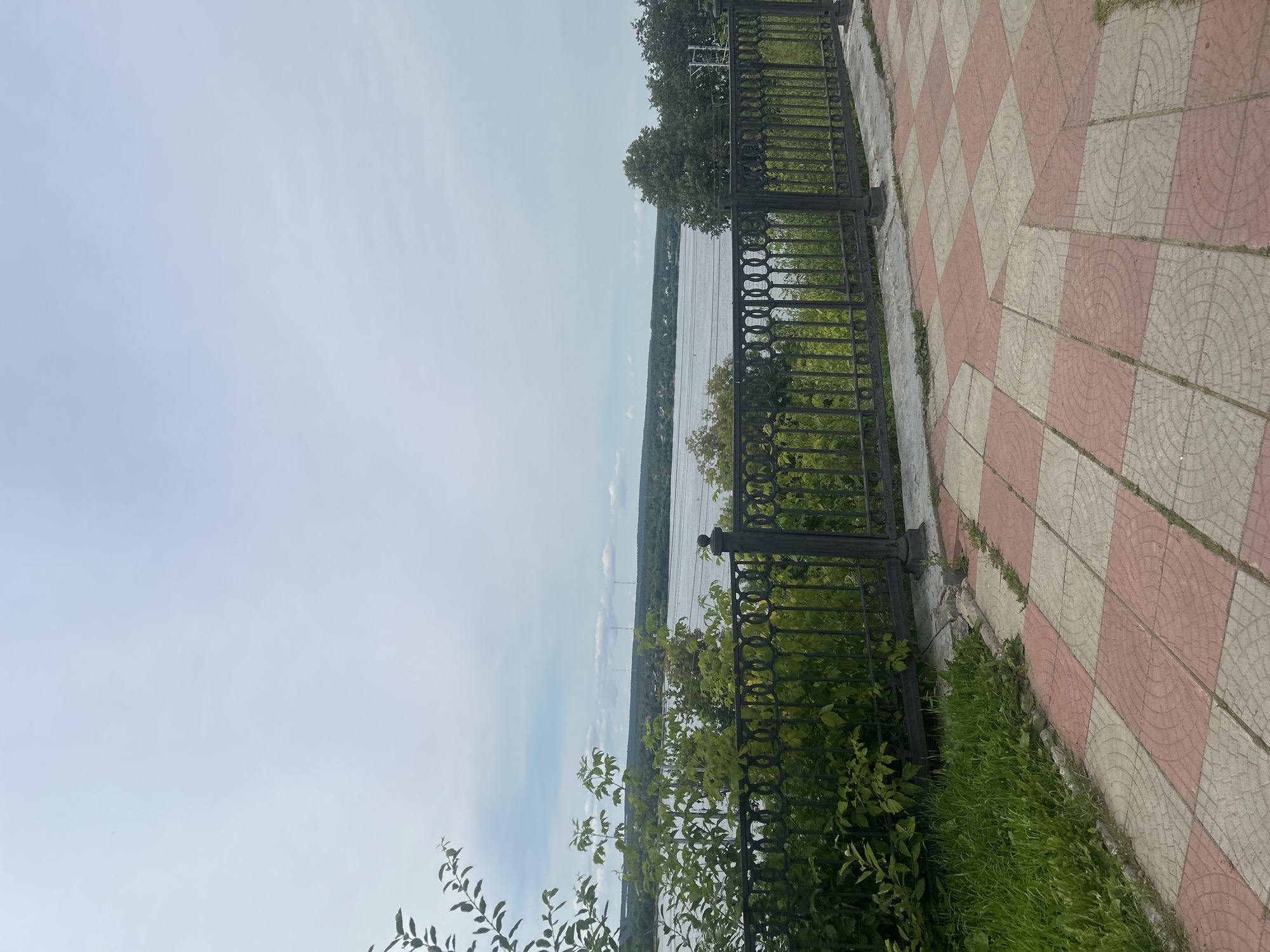 The image depicts a view from a riverside promenade in Perm, Russia. In the foreground, a patterned walkway of pink and beige square tiles leads towards a black metal railing. Beyond the railing, lush green foliage and trees frame the scene. The main subject is the wide, calm river, which stretches across the mid-ground. The opposite bank is lined with a dense forest, and a hazy horizon with scattered buildings and communication towers is visible under a cloudy, pale blue sky. The overall atmosphere is serene and overlooks a natural landscape within an urban setting.